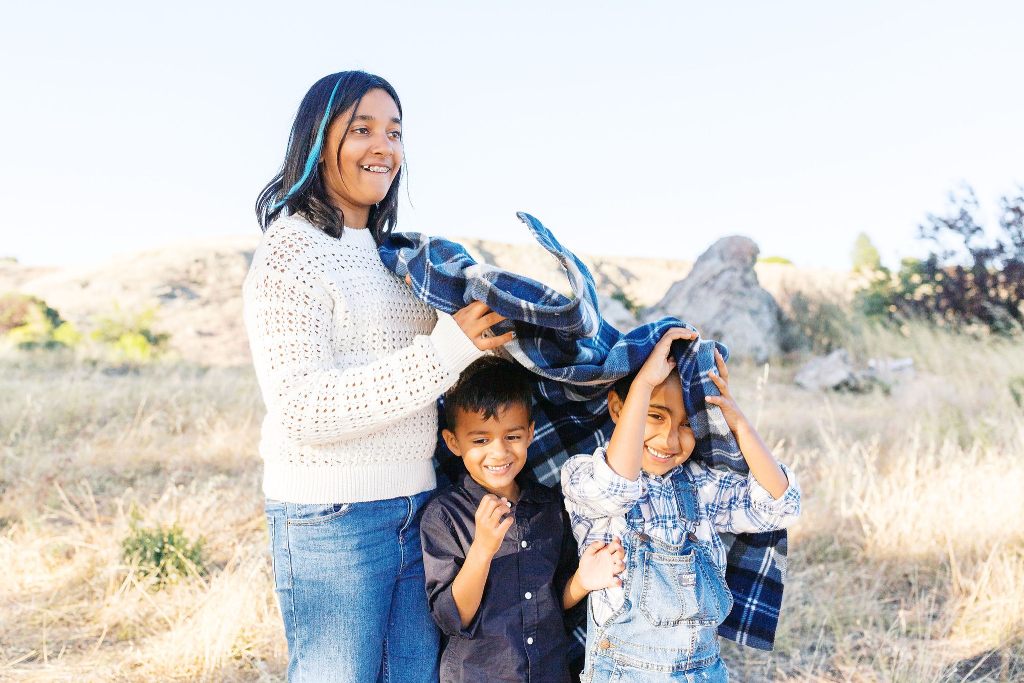 Marin family photographer captures a joyful outdoor moment as a mom playfully wraps her two laughing children in a blanket against a golden hillside backdrop — authentic lifestyle family photography in Marin County.