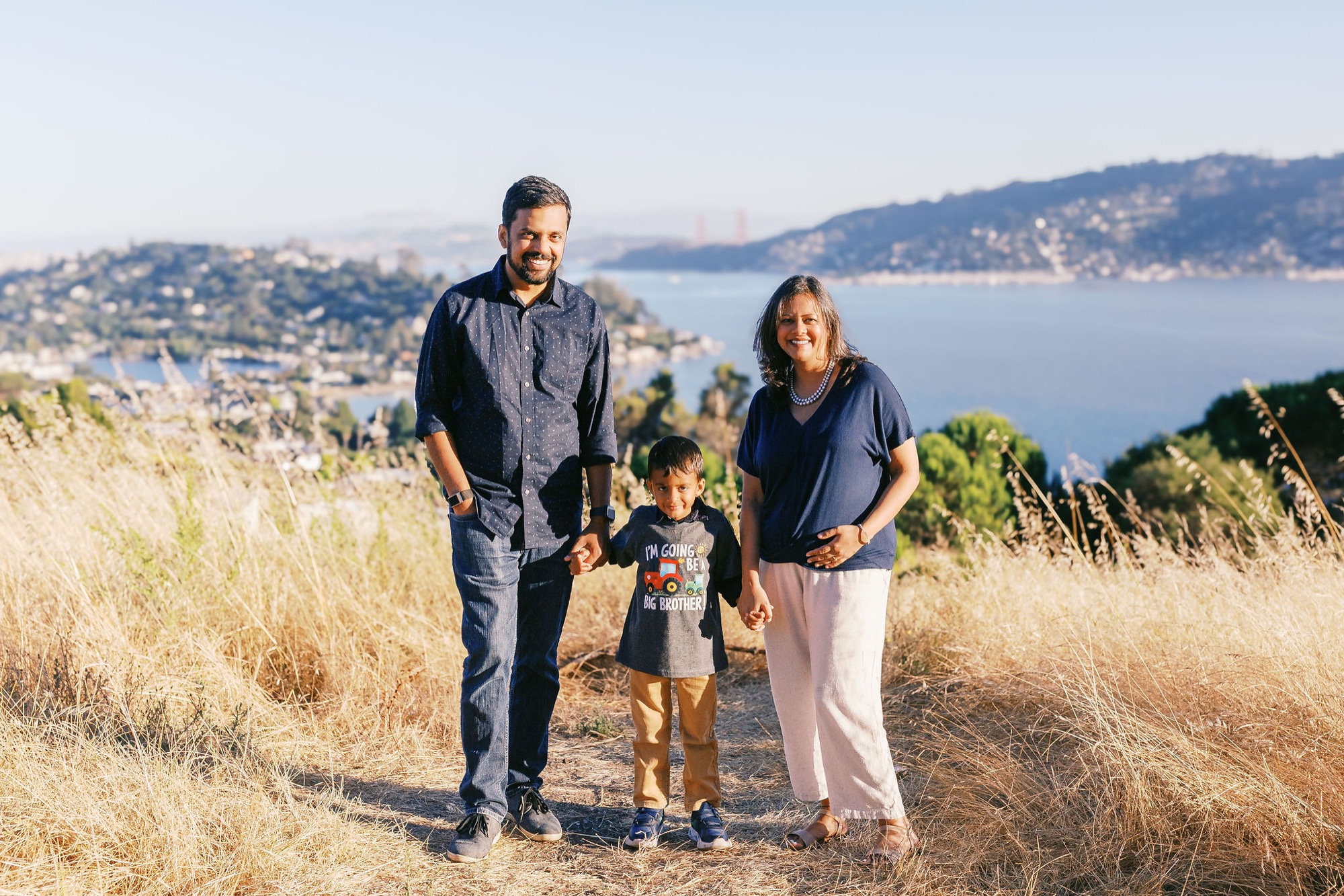 Marin family photographer captures a sweet milestone moment as a family of three holds hands on a hilltop in Marin County, the Golden Gate Bridge and San Francisco Bay glowing behind them — their little one's 'Big Brother' shirt hinting at the joy to come.