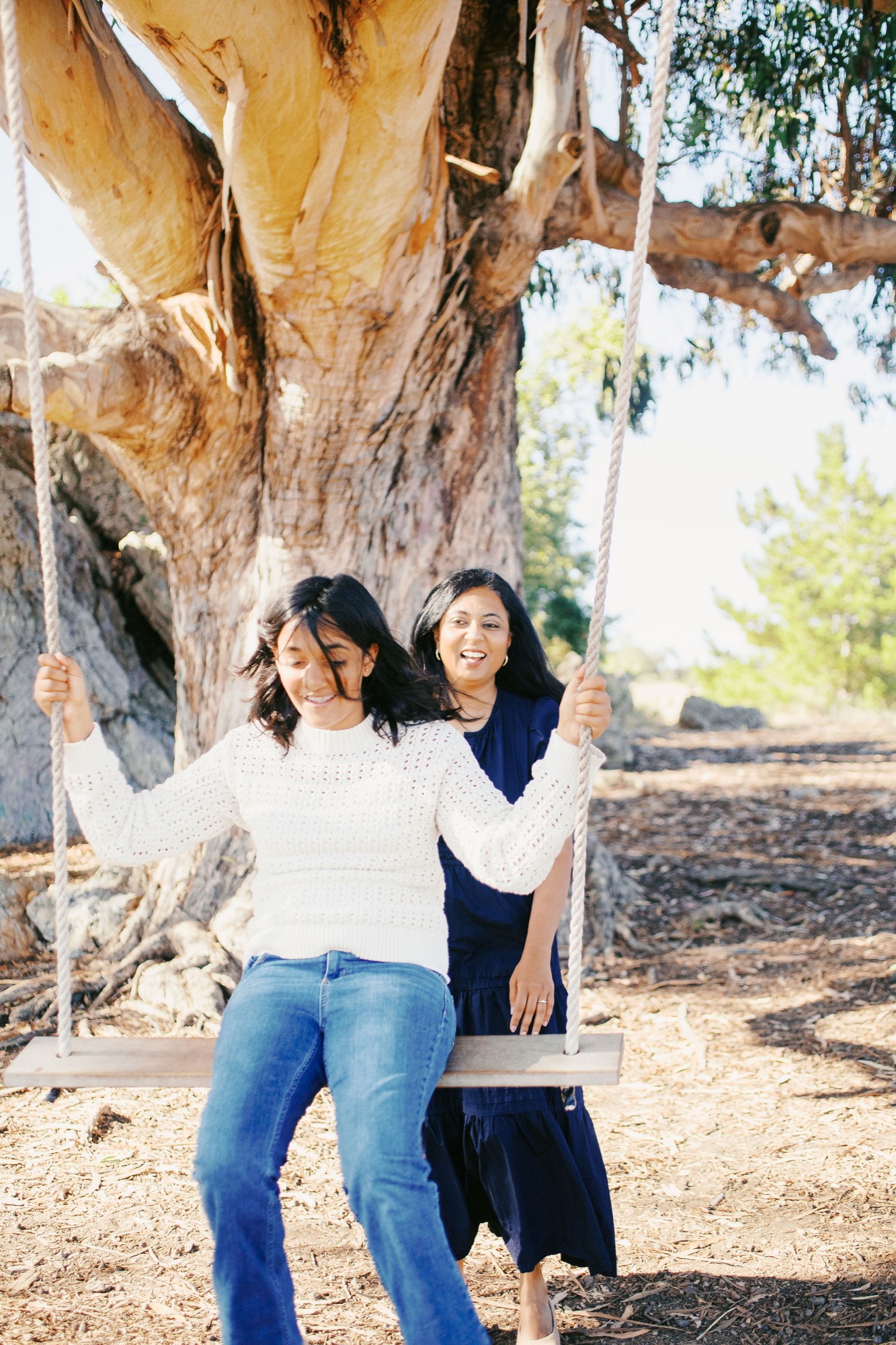 Marin family photographer captures a joyful candid moment as two women share laughter on a rope swing beneath a sprawling eucalyptus tree during an outdoor extended family session in Marin County — genuine connection and playful energy at their best.