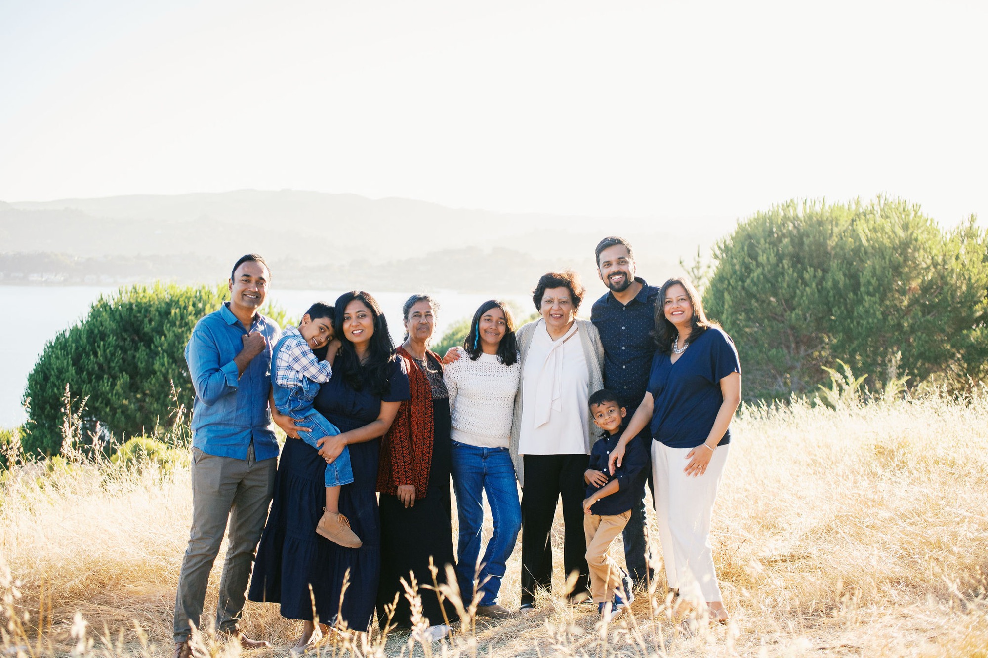 Marin family photographer captures three generations of an extended family smiling together in warm golden hour backlight at a scenic Marin County overlook, with sweeping bay views stretching out behind them — Bay Area extended family and multigenerational photography.