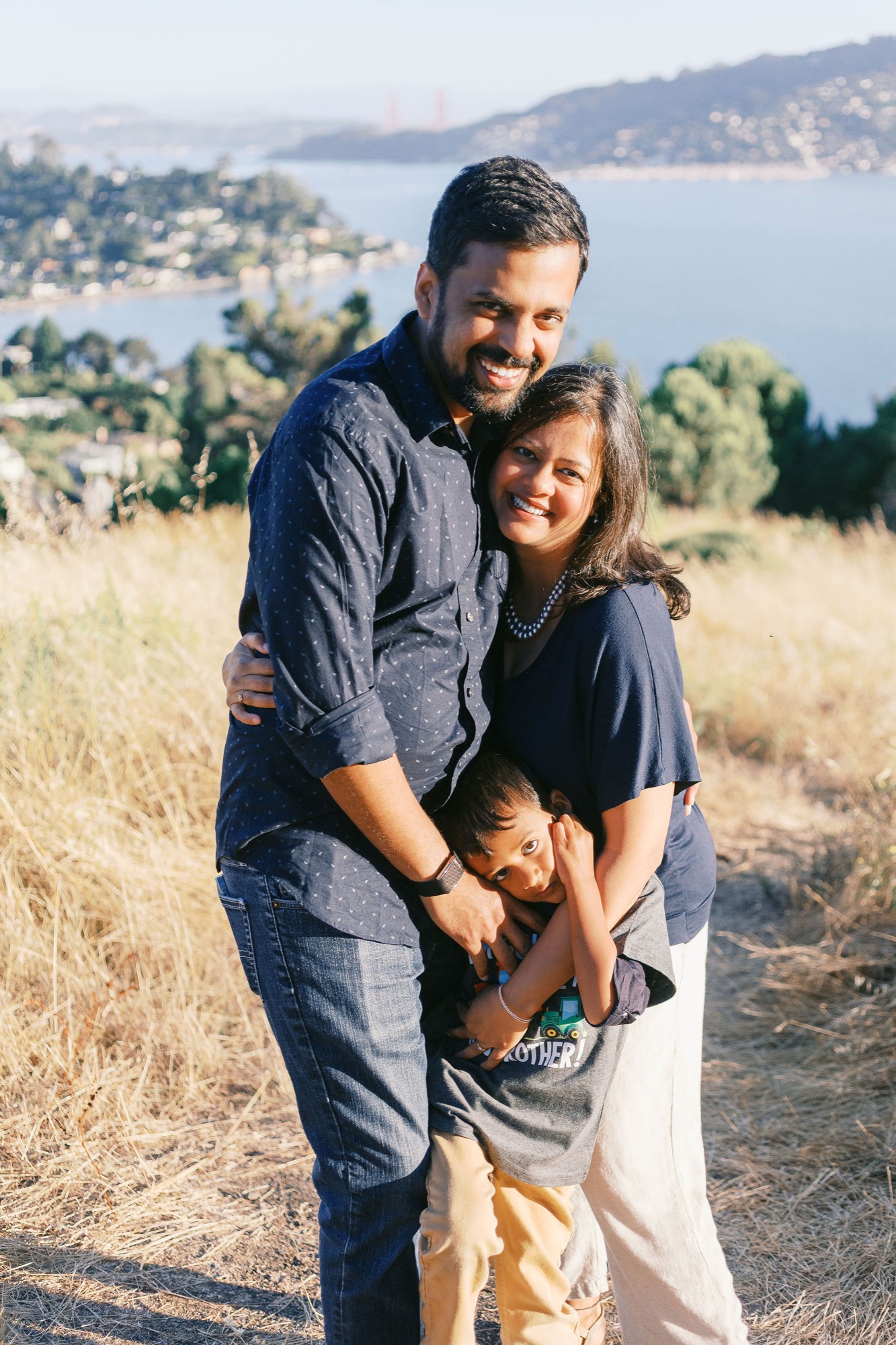 Marin family photographer captures a joyful family of three nestled together in golden afternoon light on a Marin hillside, with the San Francisco Bay and Golden Gate Bridge glowing softly in the distance — outdoor family portrait photography in Marin County.