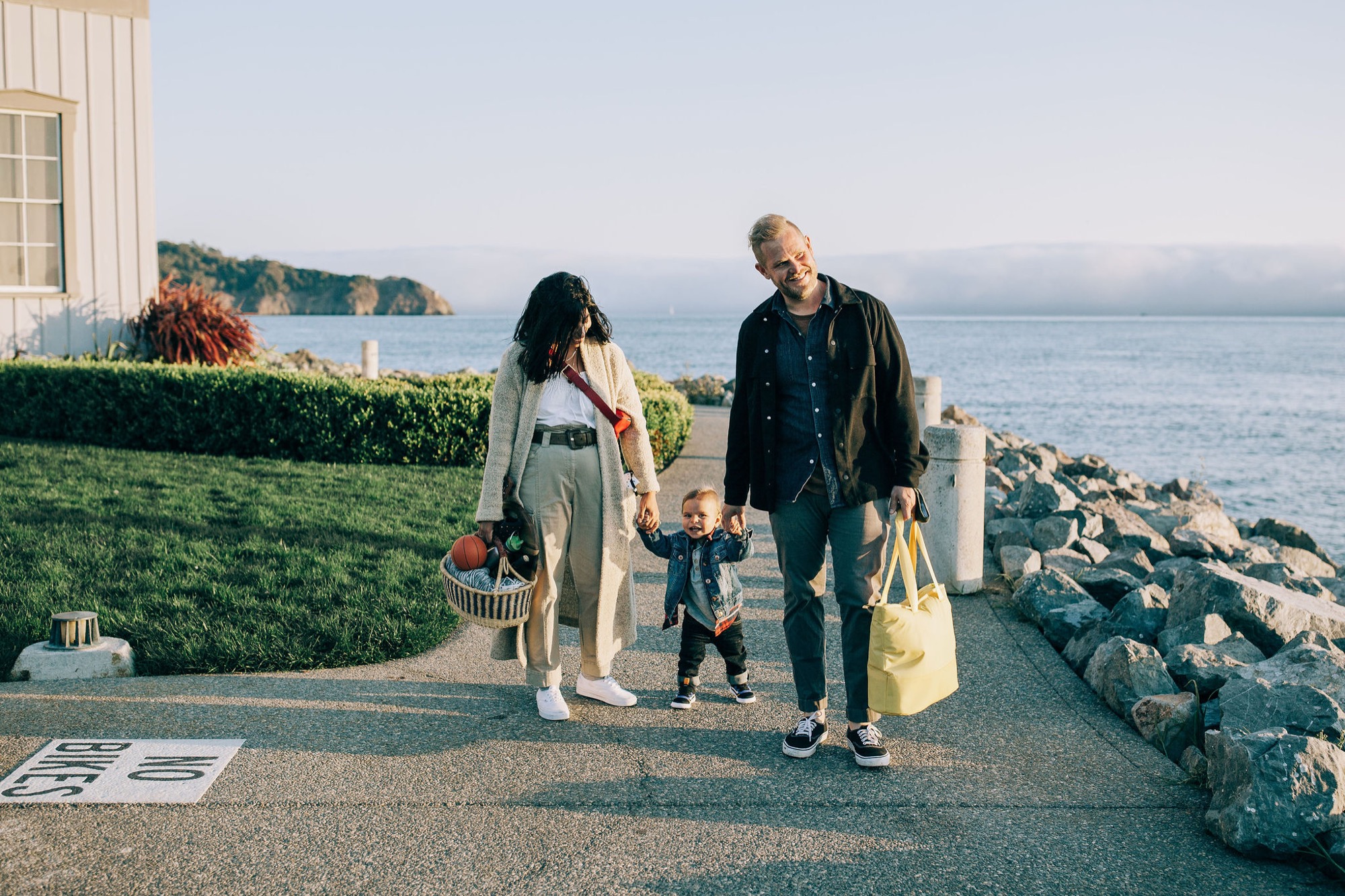 A family of three strolls along a coastal pathway at sunset, the child between them holding both parents' hands while the bay stretches behind them
