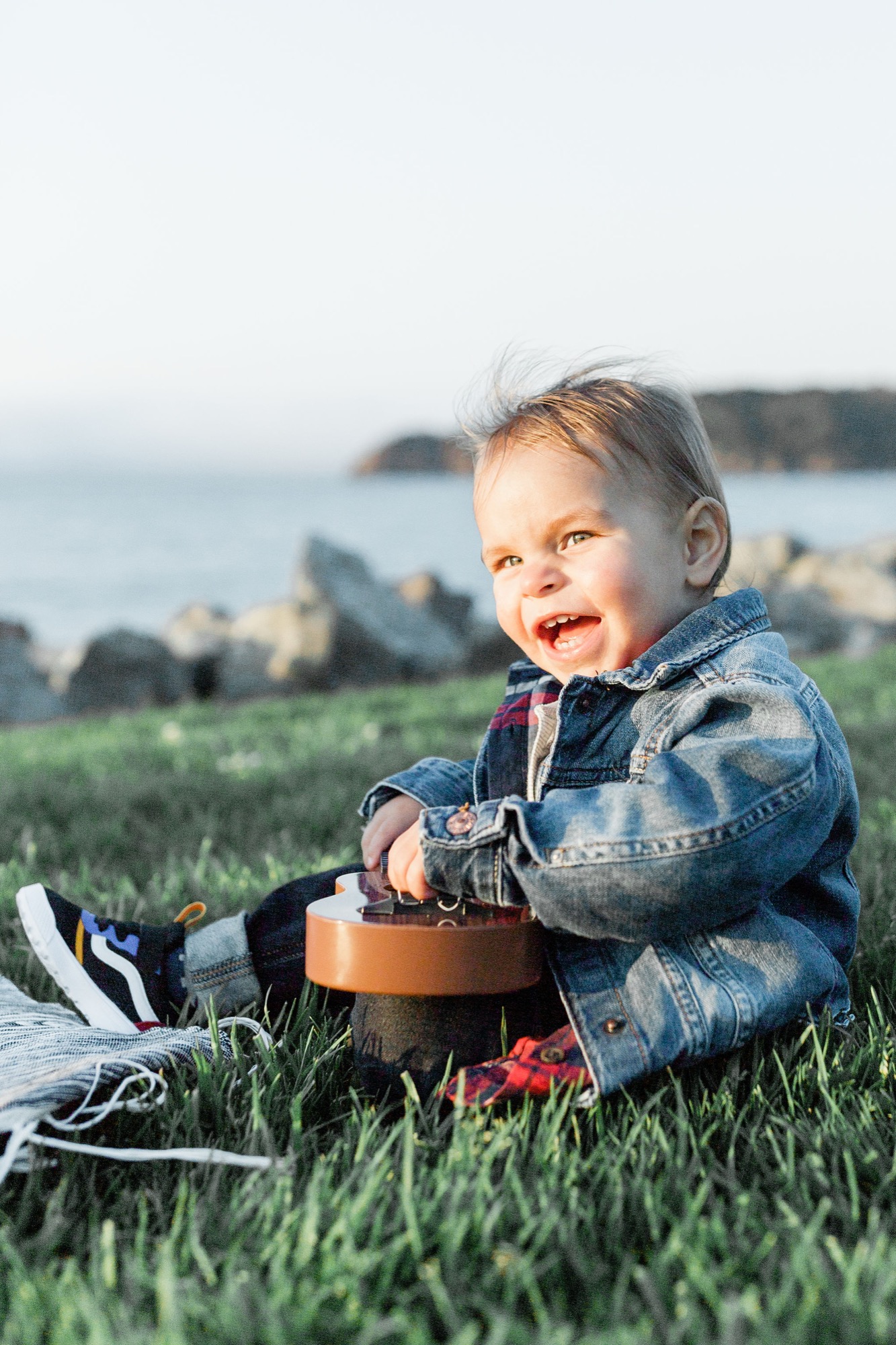 Toddler in denim jacket sits on grass with ocean cliffs behind, smiling at camera in golden afternoon light