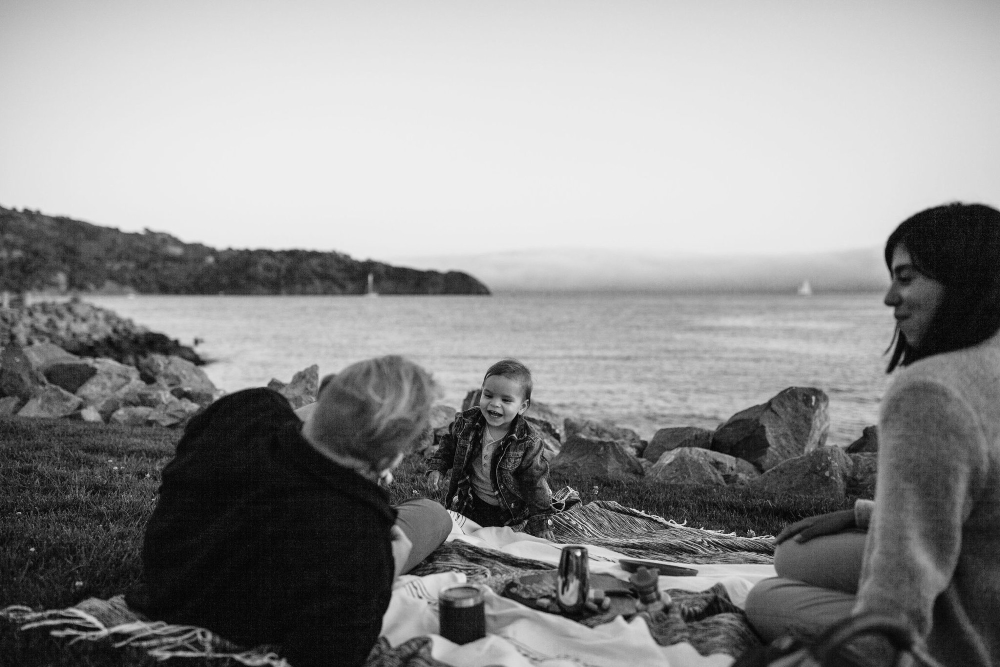 Four people gathered on rocky beach at Tiburon Waterfront, sharing a meal with calm bay waters and forested hills behind them