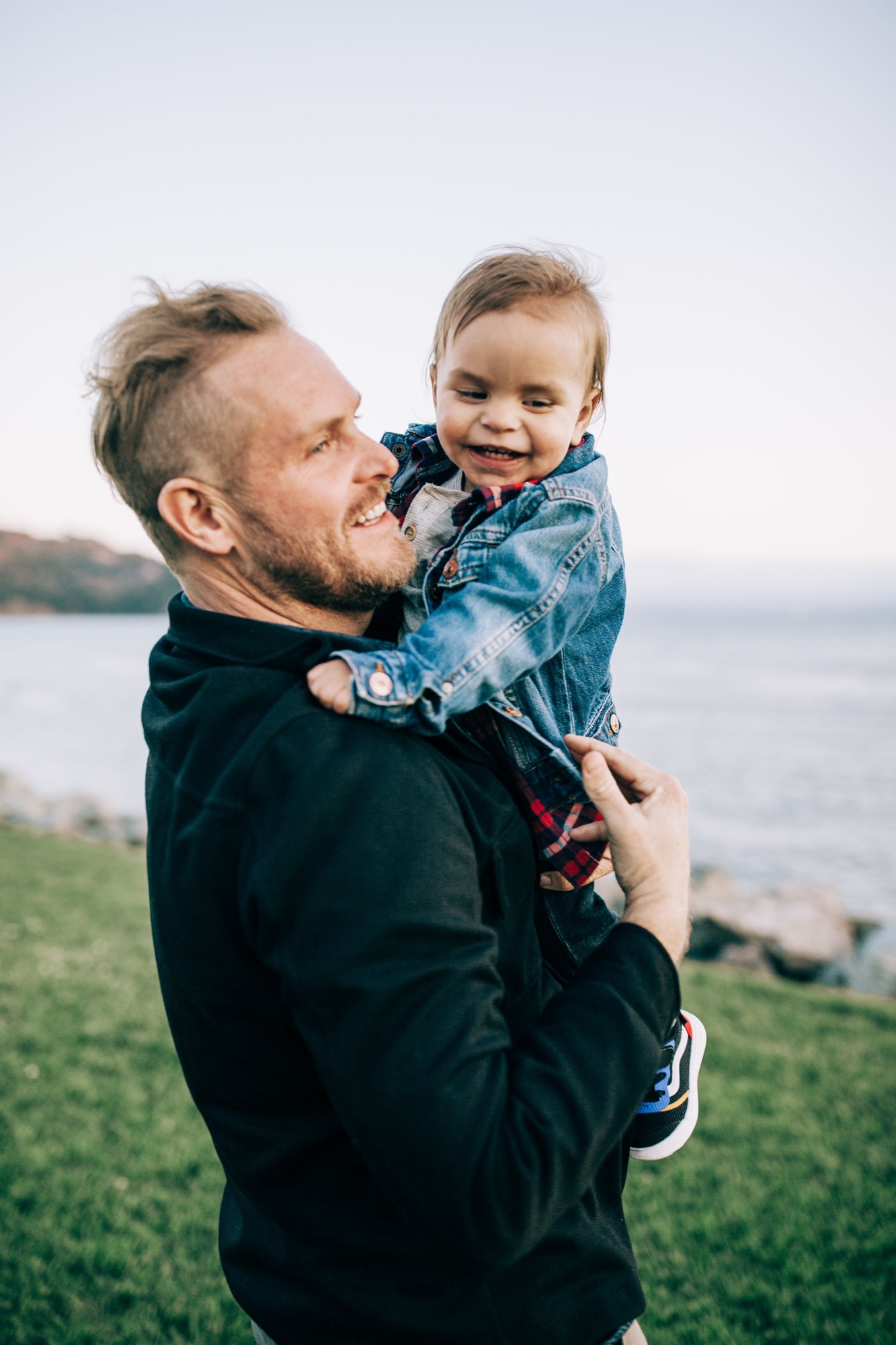 A bearded father in dark jacket holds his smiling toddler against a soft-focused waterfront backdrop with grassy terrain