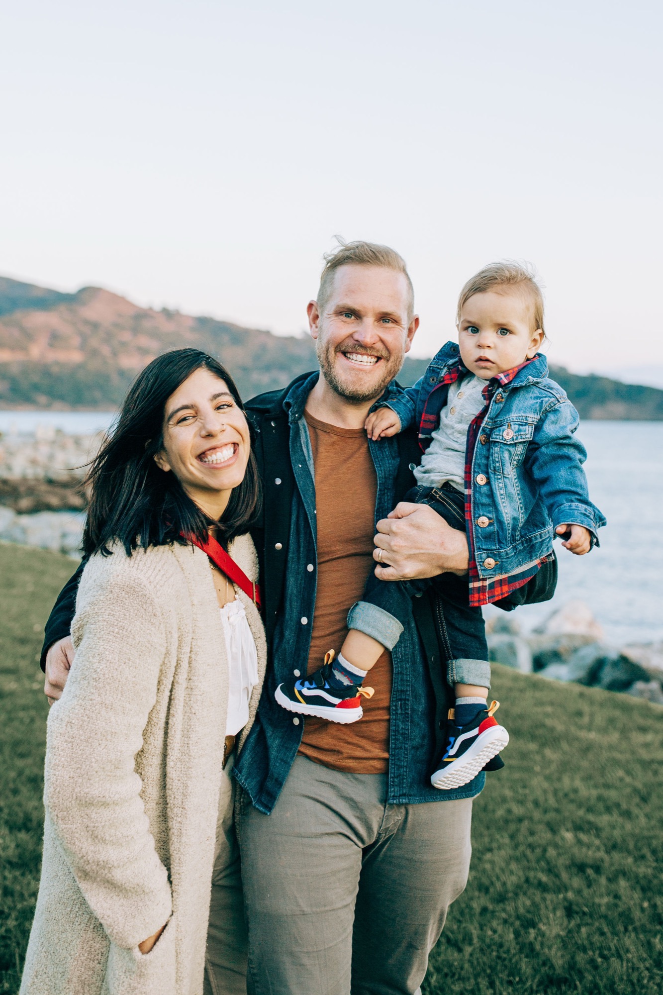 A smiling father holds his young son in a denim jacket while the mother leans in, all three positioned against a scenic waterfront backdrop with rolling hills