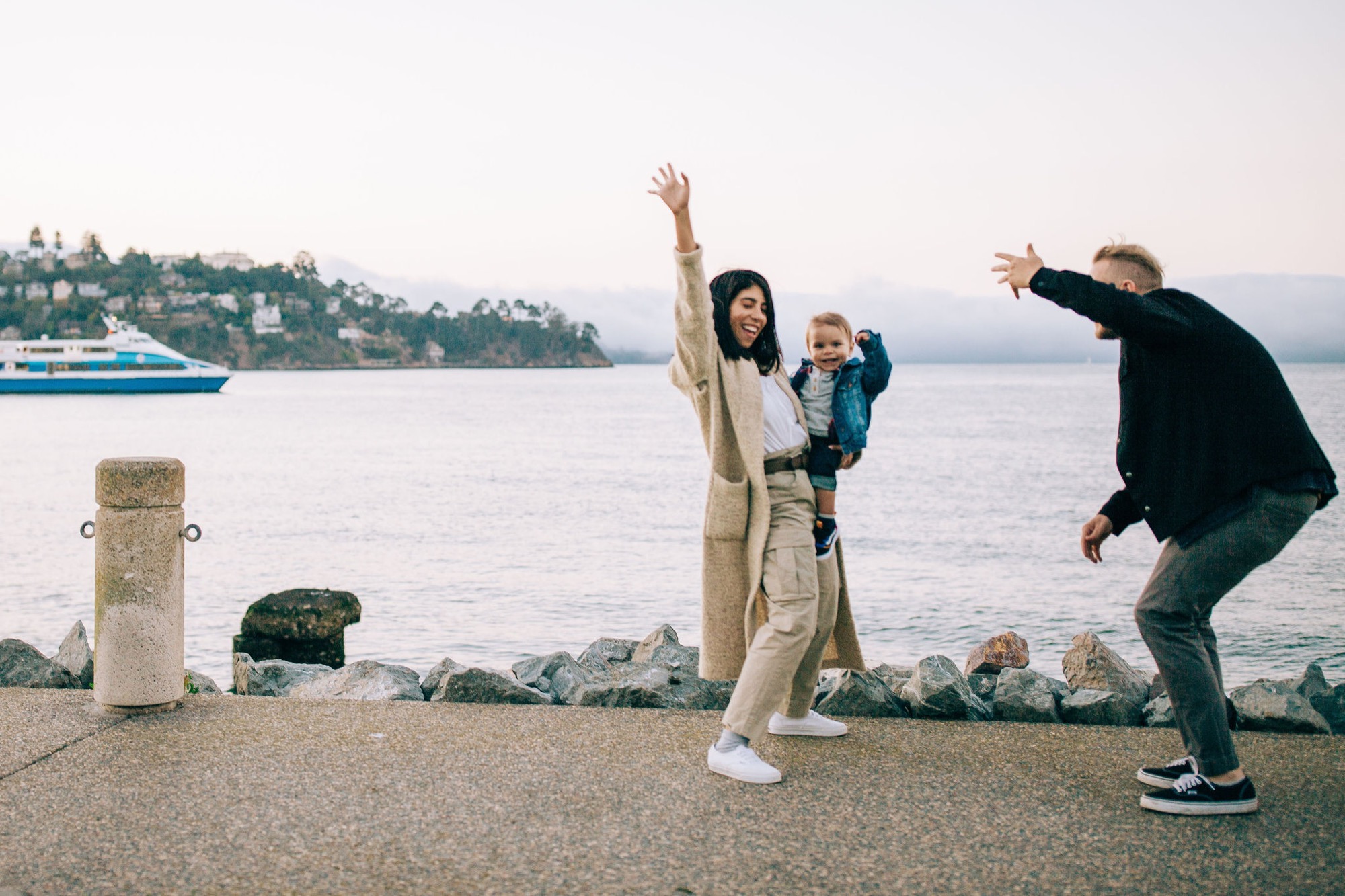 Family playfully poses on sandy beach with woman's arms raised high, man crouching beside her, child between them, ferry visible across calm water