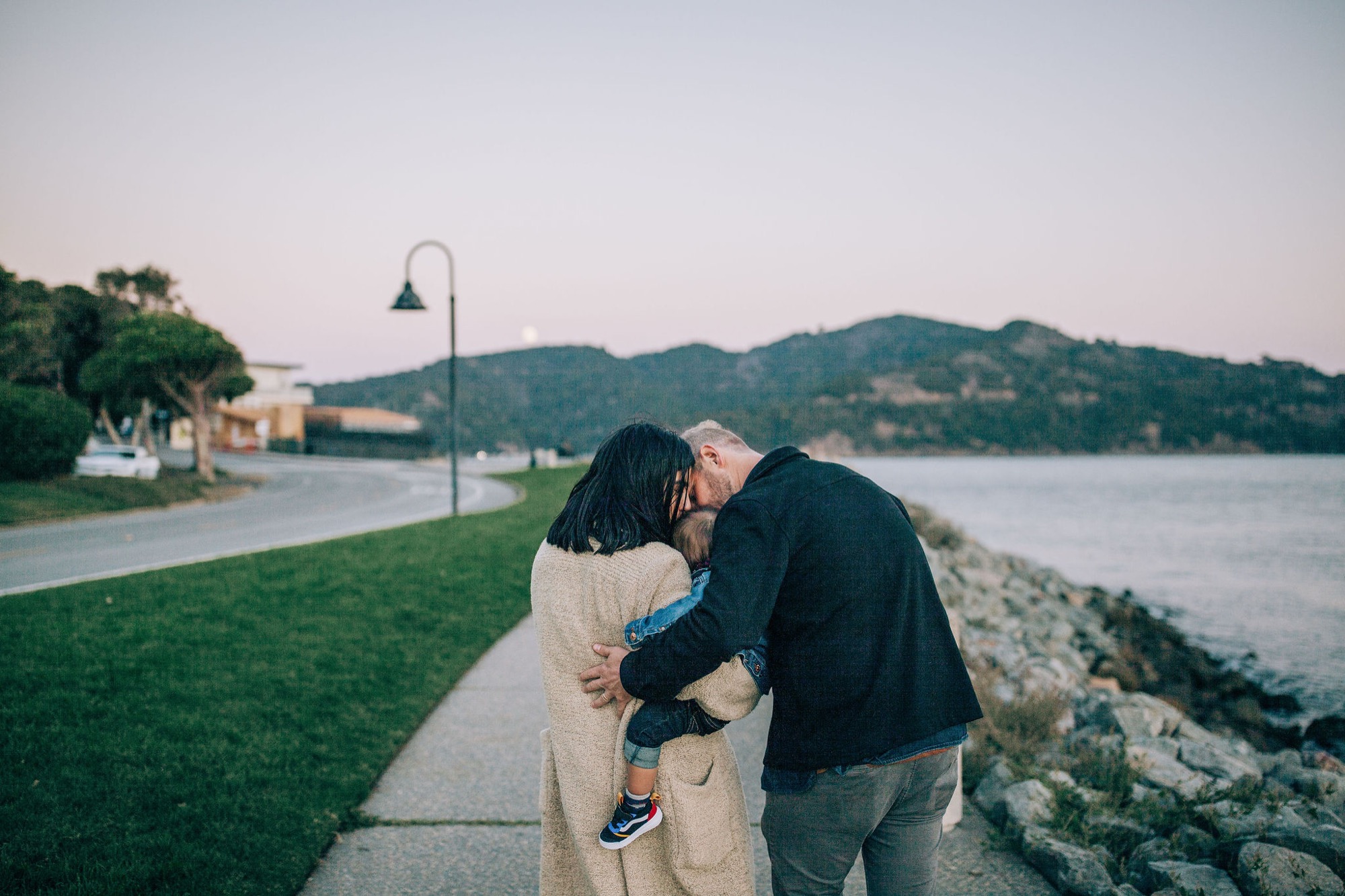 A couple embraces on a waterfront path at dusk, with forested hills and calm water reflecting the twilight sky behind them