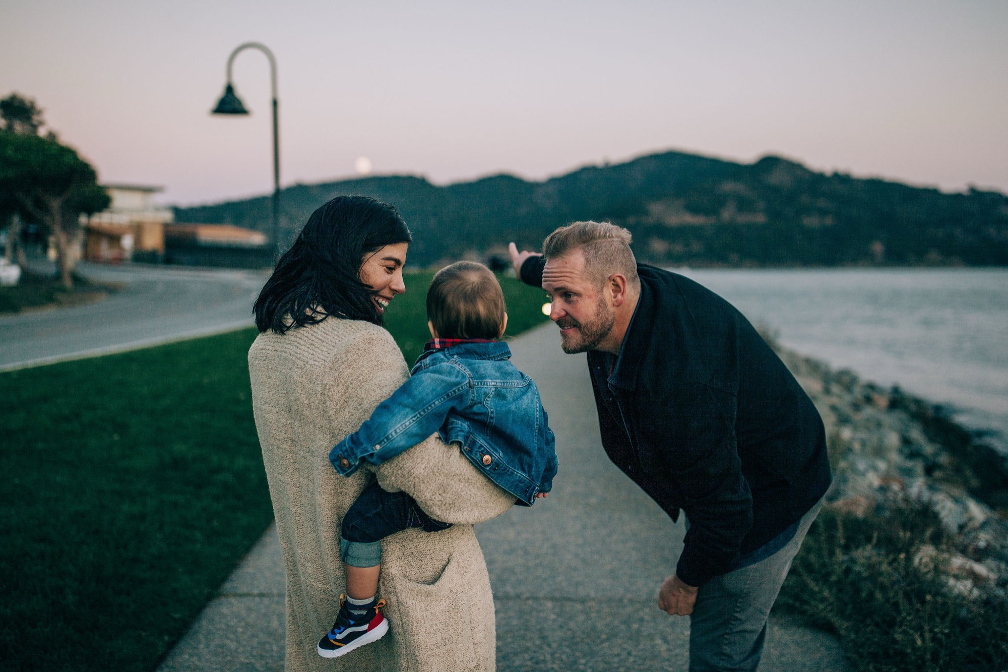 Parents crouch beside their young child at the waterfront as golden-hour light bathes the rocky shoreline and distant hills