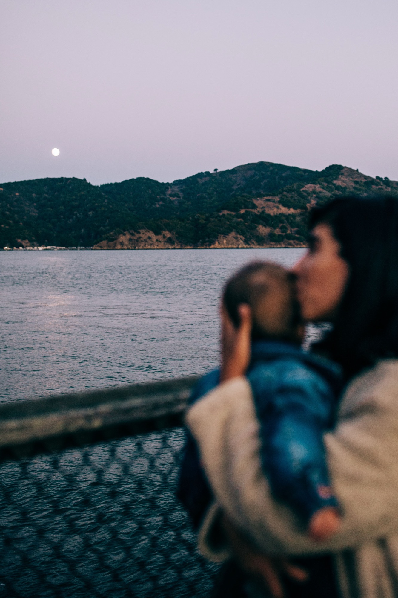 A woman holds her child while gazing toward moonlit hills across calm bay waters at dusk