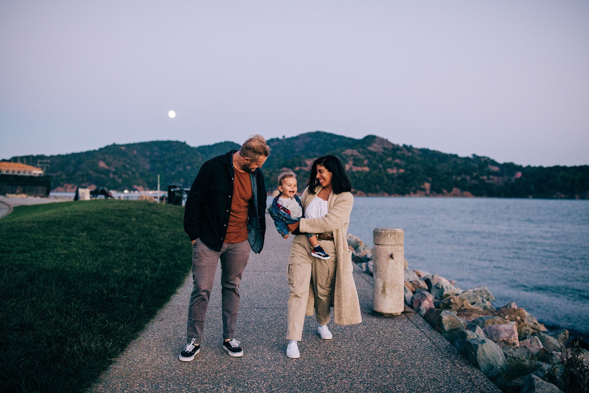 Family of three stands on a waterfront path at dusk, with the father leaning toward his young son while the mother smiles nearby, the moon rising over forested hills behind them