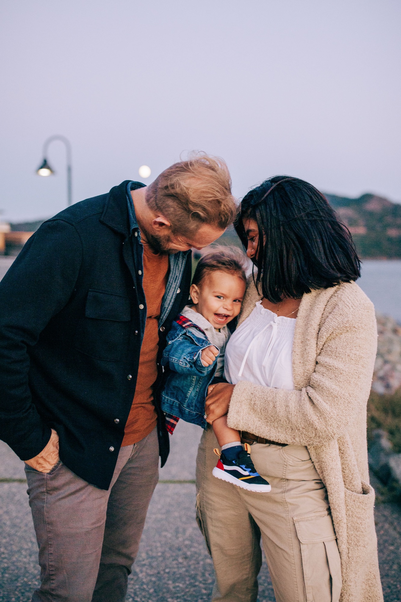 Two adults lean in to kiss a toddler between them on a waterfront path at golden hour, their warm expressions radiating affection