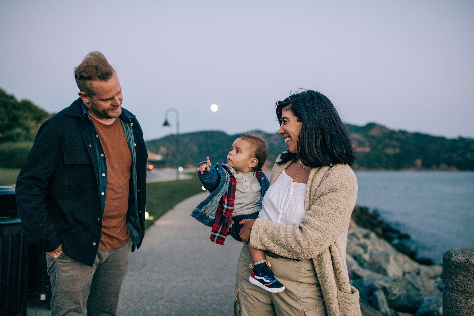 A young family laughs together on a waterfront path at dusk, with the moon rising over the bay behind them