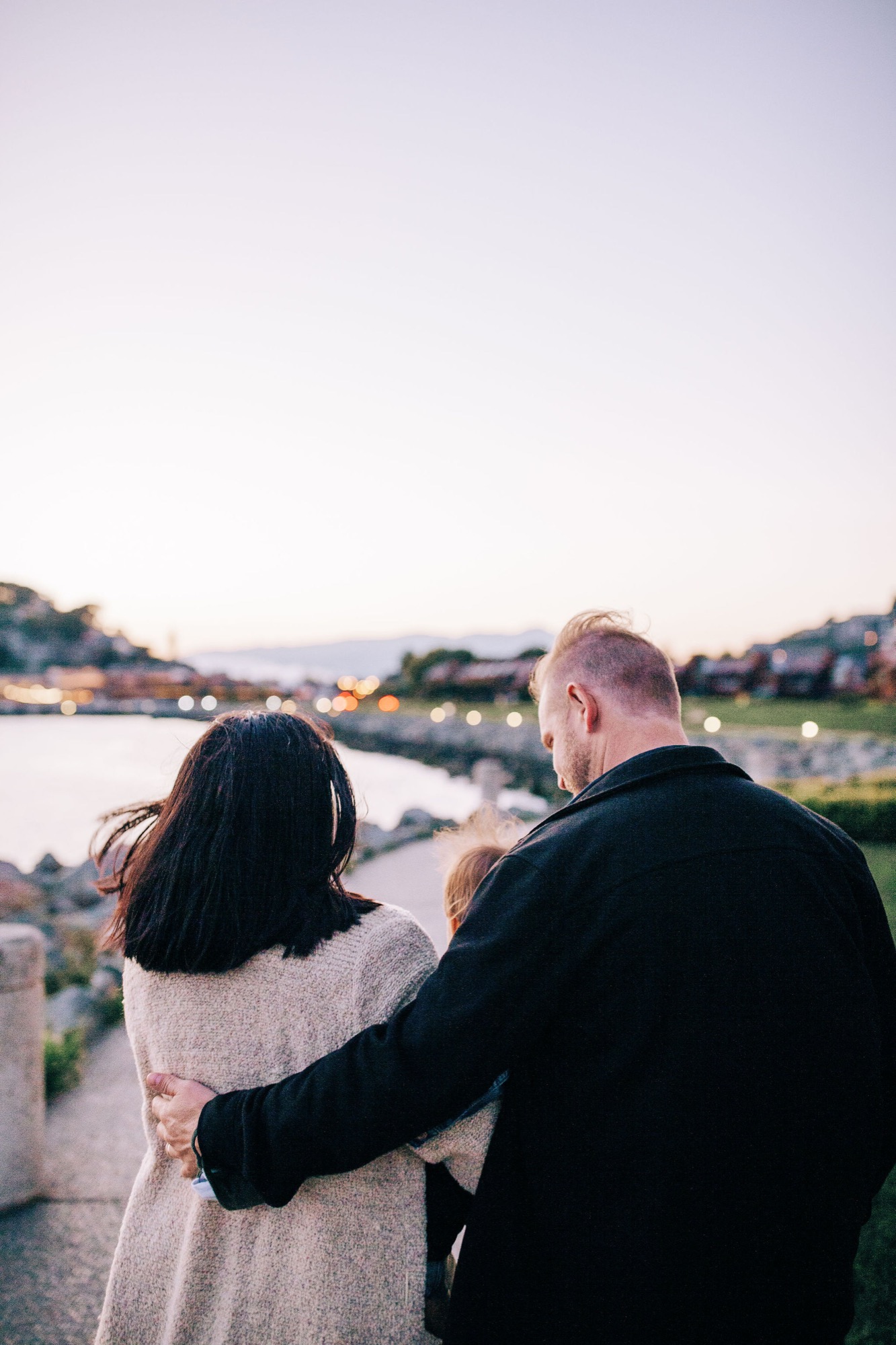 Couple gazes toward the waterfront at golden hour, his arm around her shoulders as distant hills glow in soft evening light