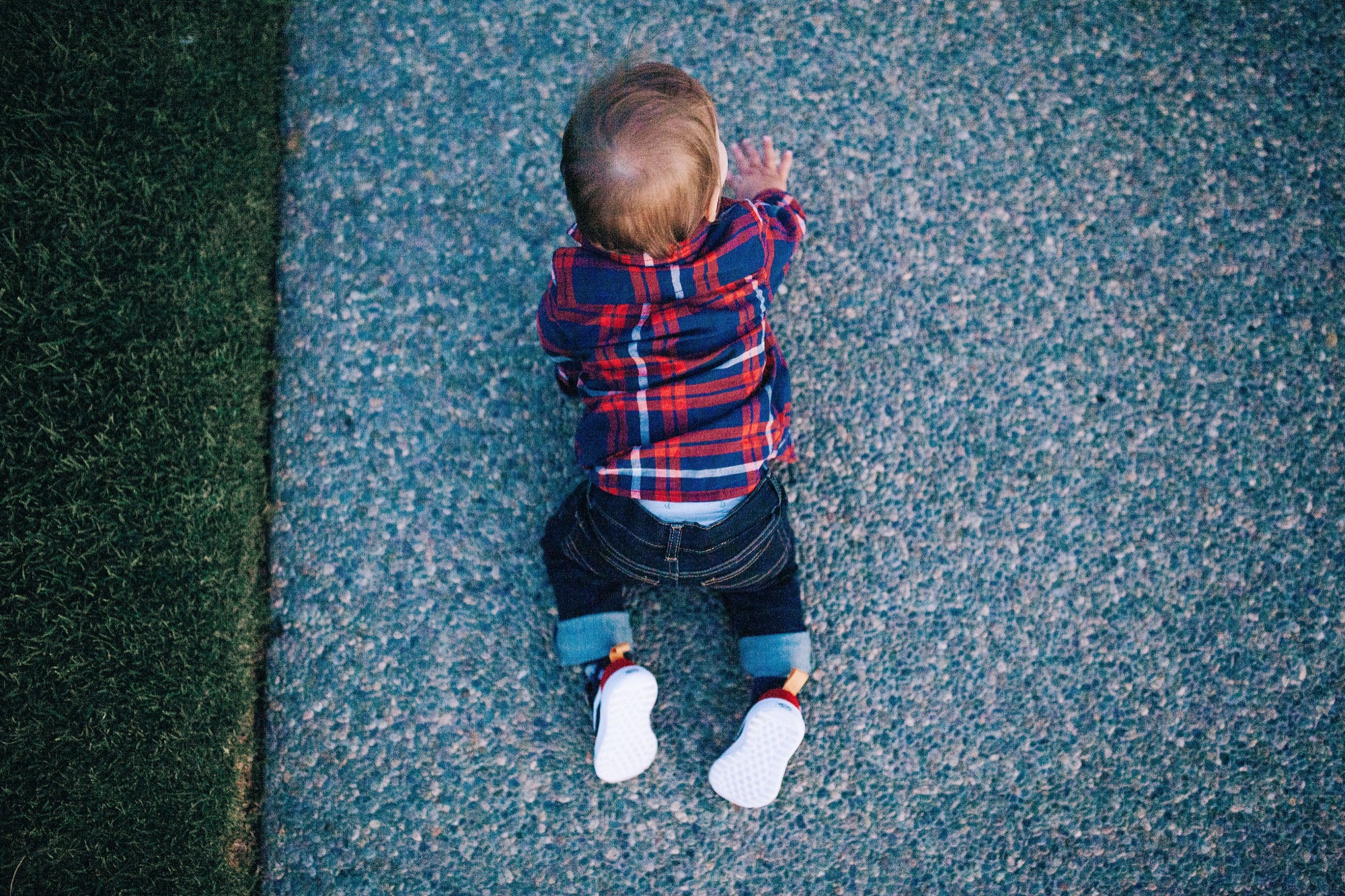 Toddler in red plaid shirt lies on textured ground, wearing white socks, captured from directly above in natural daylight