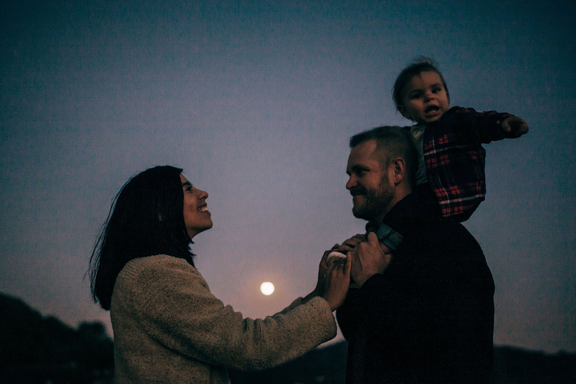 A family silhouetted against a twilight sky and moon, with a father holding his child on his shoulders while the mother gazes upward beside them