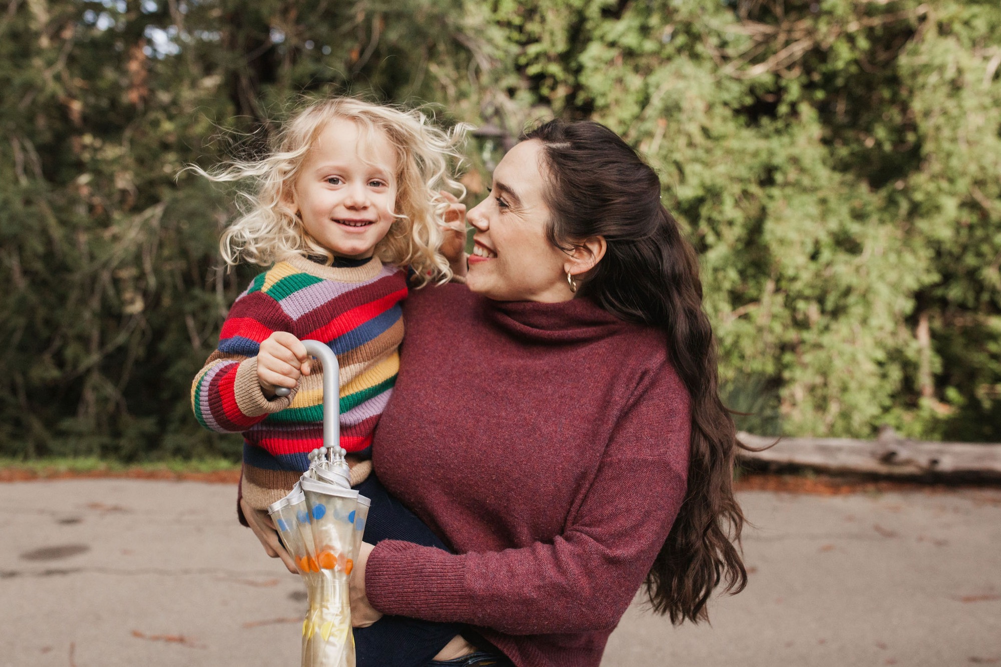 A woman in a burgundy turtleneck holds her laughing daughter wearing a colorful striped sweater, both beaming at each other with genuine joy against evergreen trees