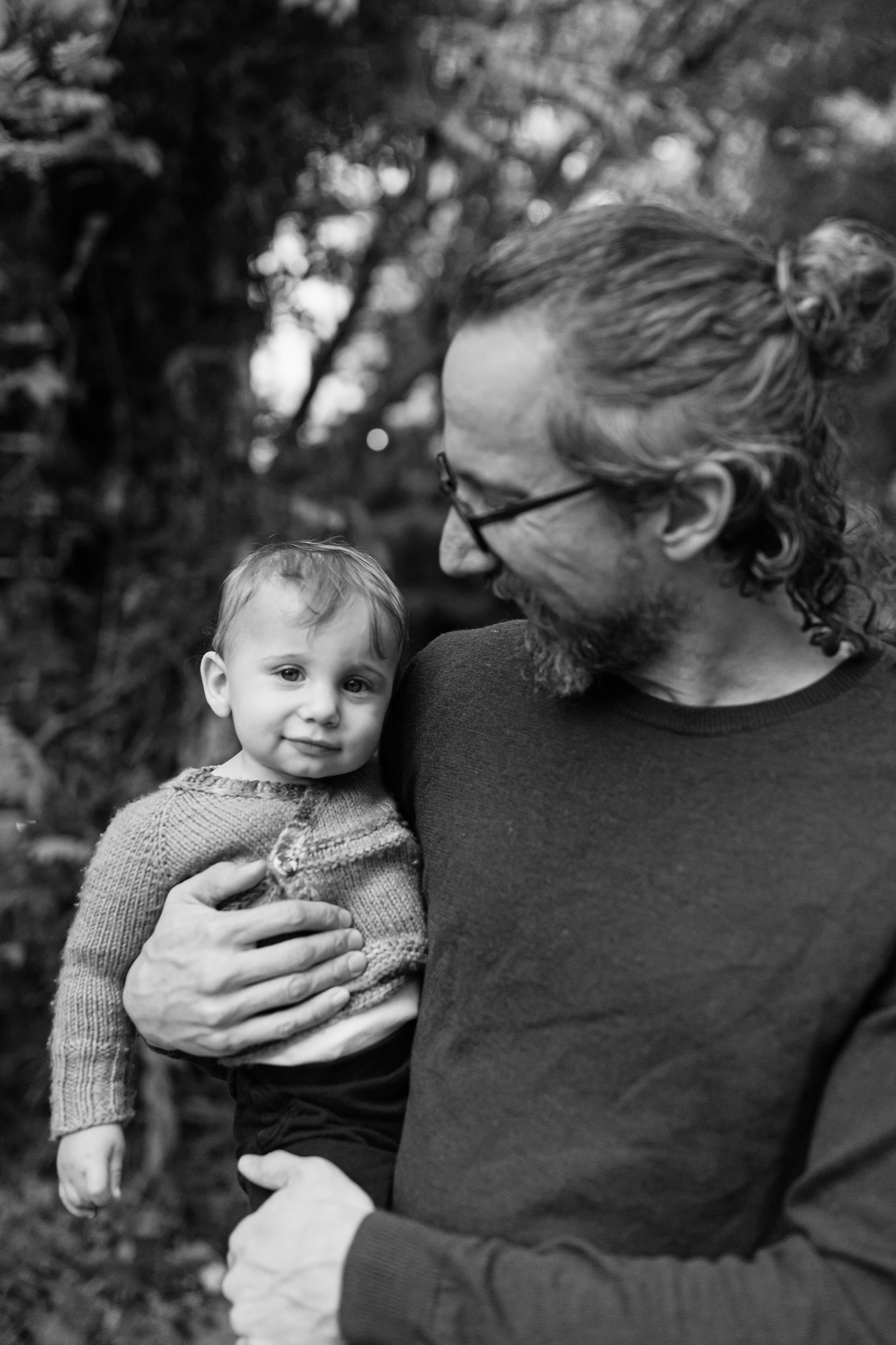 A bearded man in glasses holds his young son against his chest, both gazing thoughtfully away as dappled sunlight filters through trees behind them