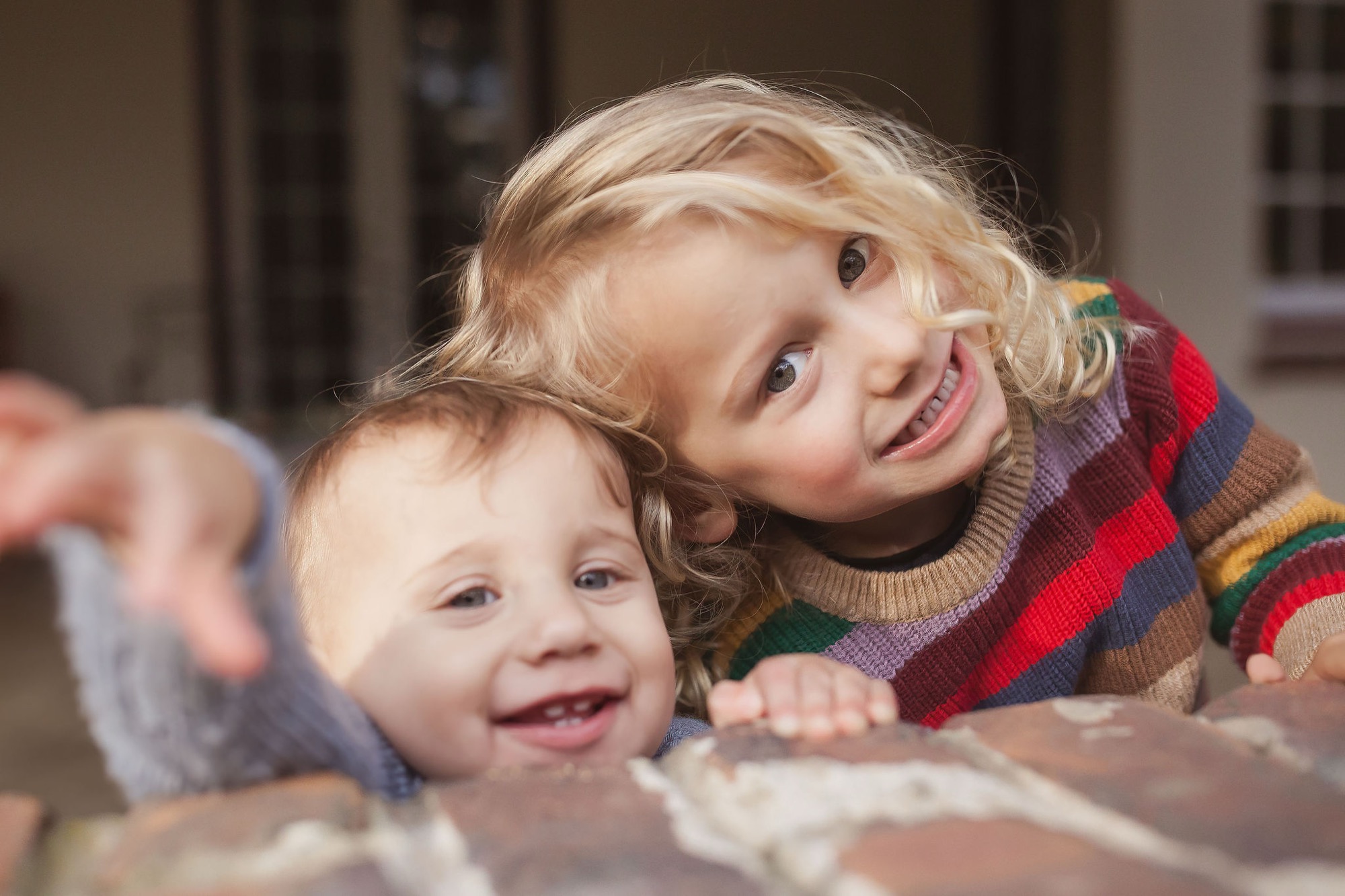Two young children beam with genuine joy, their blonde curls catching warm sunlight as they lean together against weathered stone