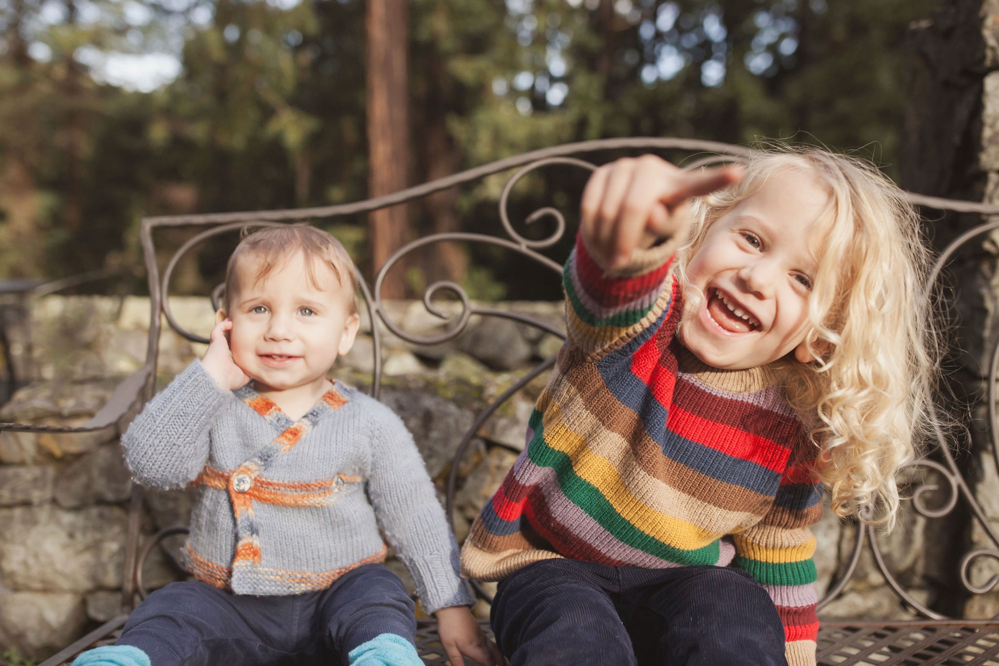 Two young children sit together on an ornate iron bench, the older girl laughing joyfully while the toddler smiles softly in warm afternoon sunlight