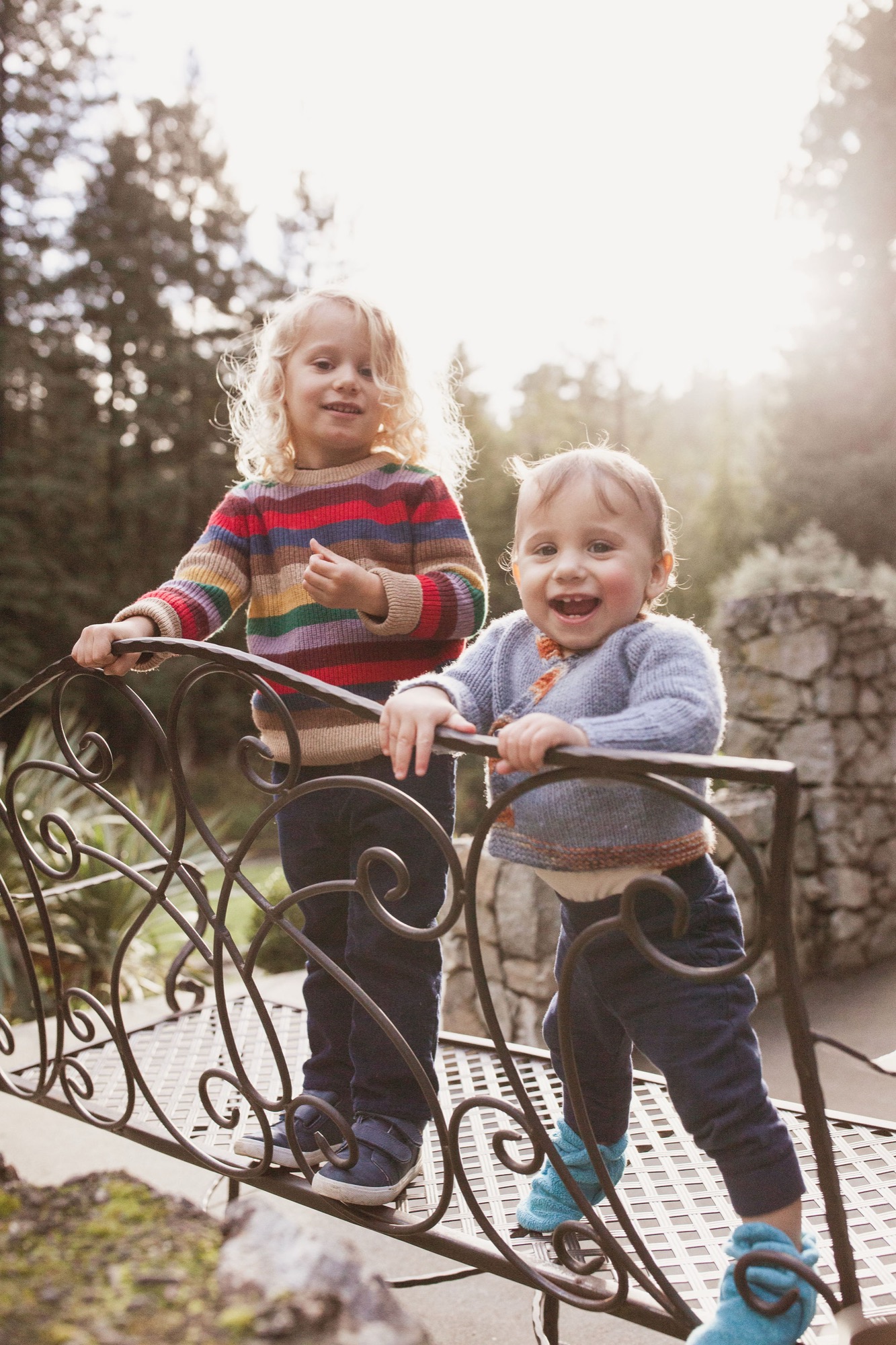 Two toddlers with blonde hair lean against ornate iron railings, beaming with genuine joy as golden sunlight filters through coastal redwoods behind them