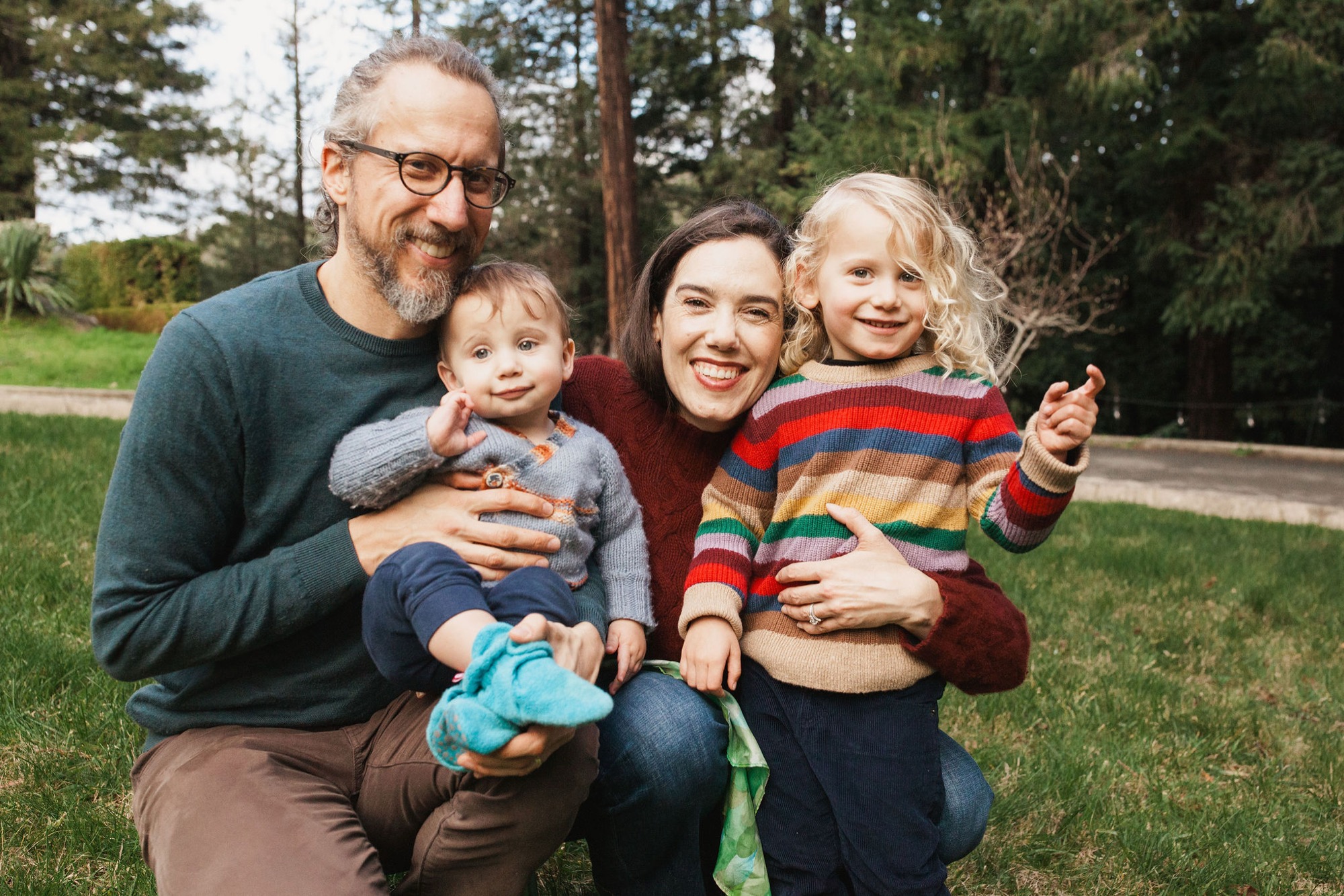 A family of four crouches together on a sunlit lawn, with the father and mother beaming while holding their two young children in colorful knit sweaters