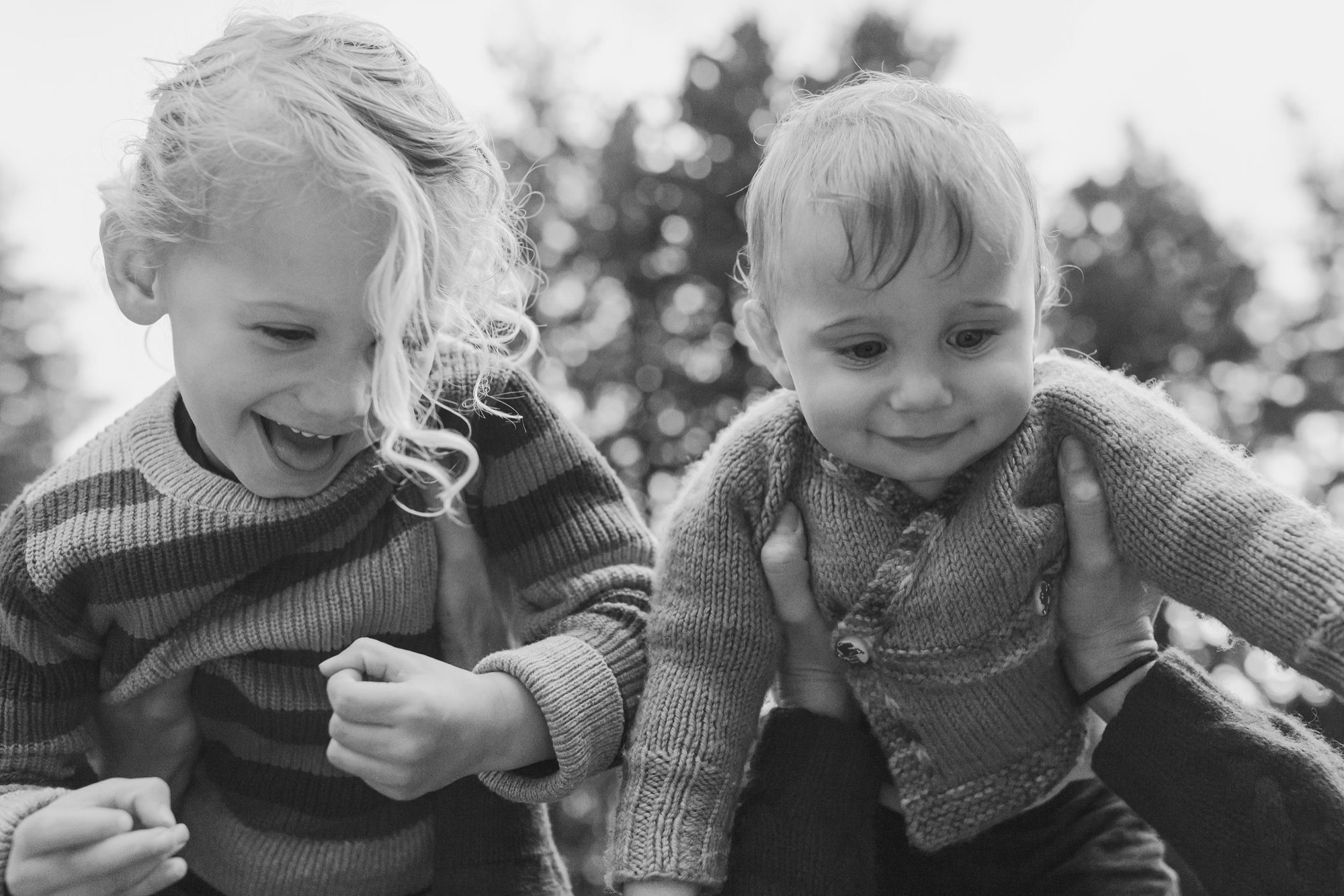 Two young children in knit sweaters laugh together outdoors, their genuine joy captured in soft natural light with blurred greenery behind
