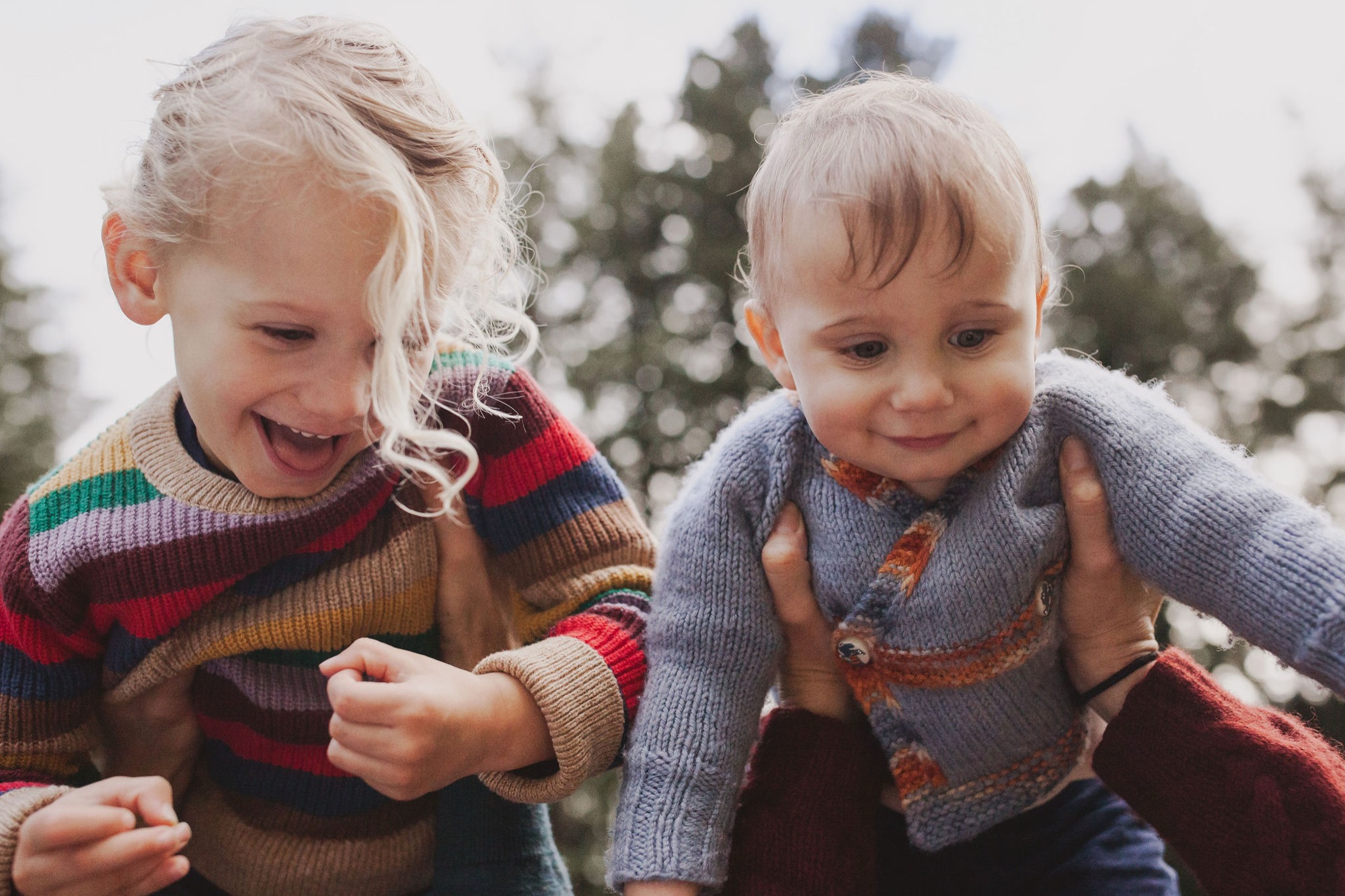 Two blonde toddlers in colorful striped cardigans laugh joyfully outdoors, their genuine expressions and animated body language capturing pure childhood delight