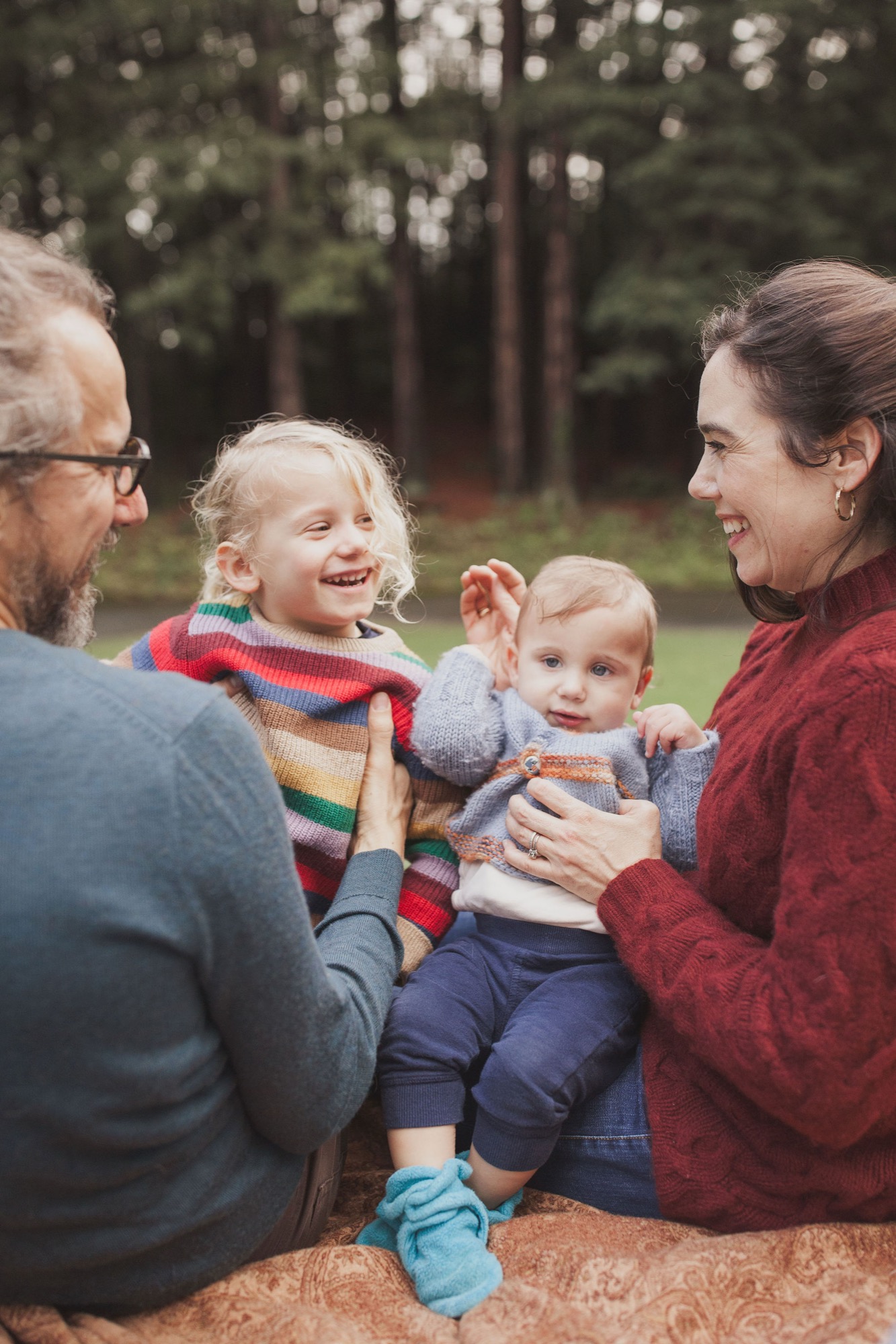 A bearded father in glasses and mother in burgundy sweater laugh with their two young children seated on a blanket, surrounded by towering redwoods