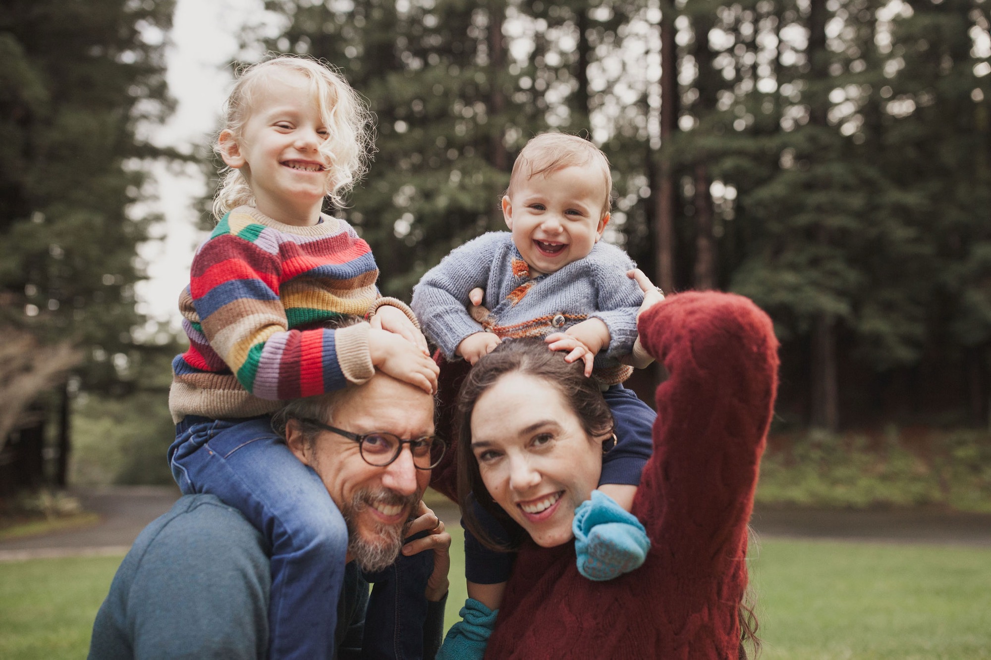 Two smiling parents carry their young children on shoulders in a sunlit forest clearing, all beaming with genuine joy