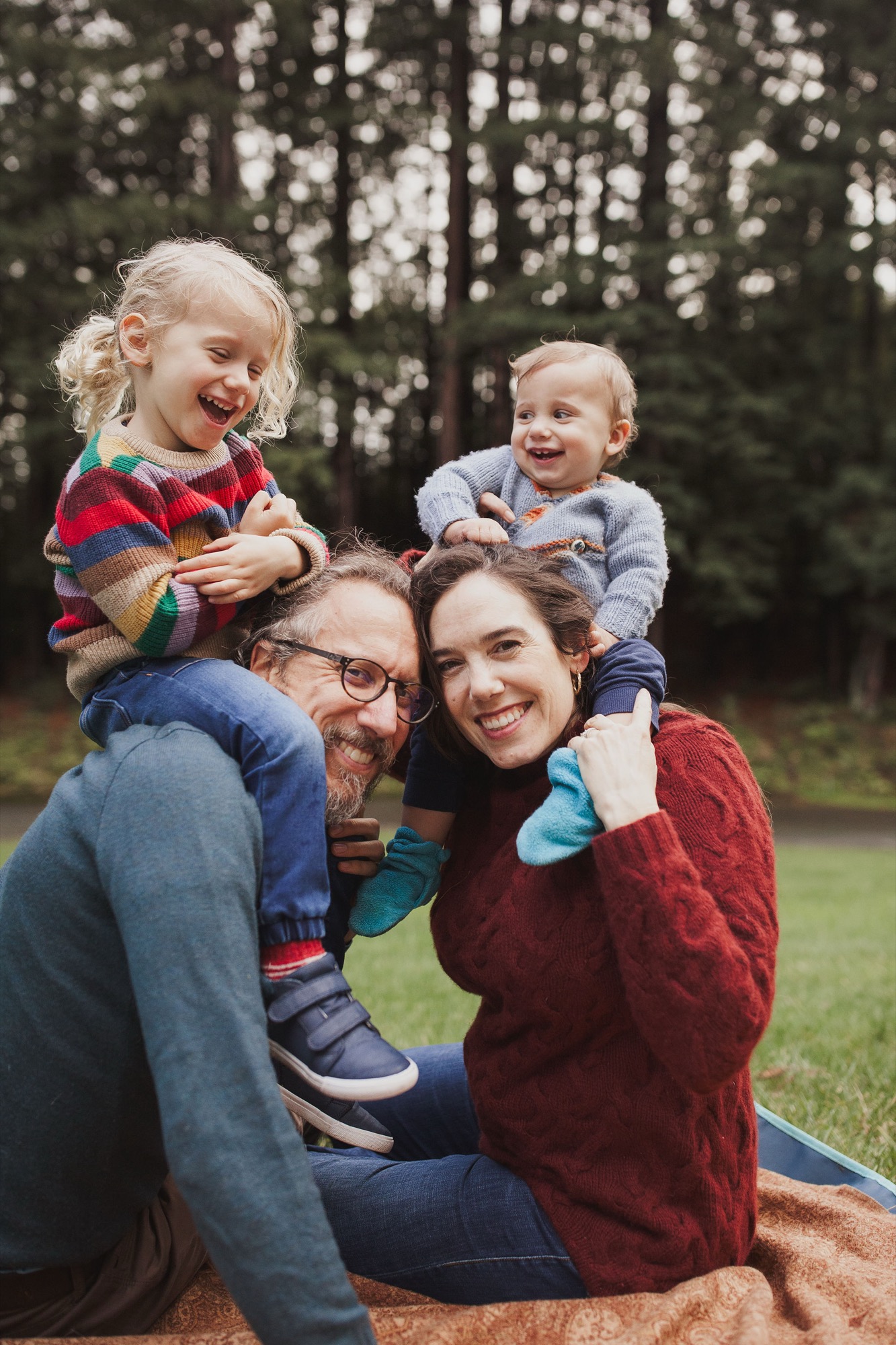 Two adults kneel on a blanket with two young children on their shoulders, all beaming with genuine joy in a sunlit forest clearing