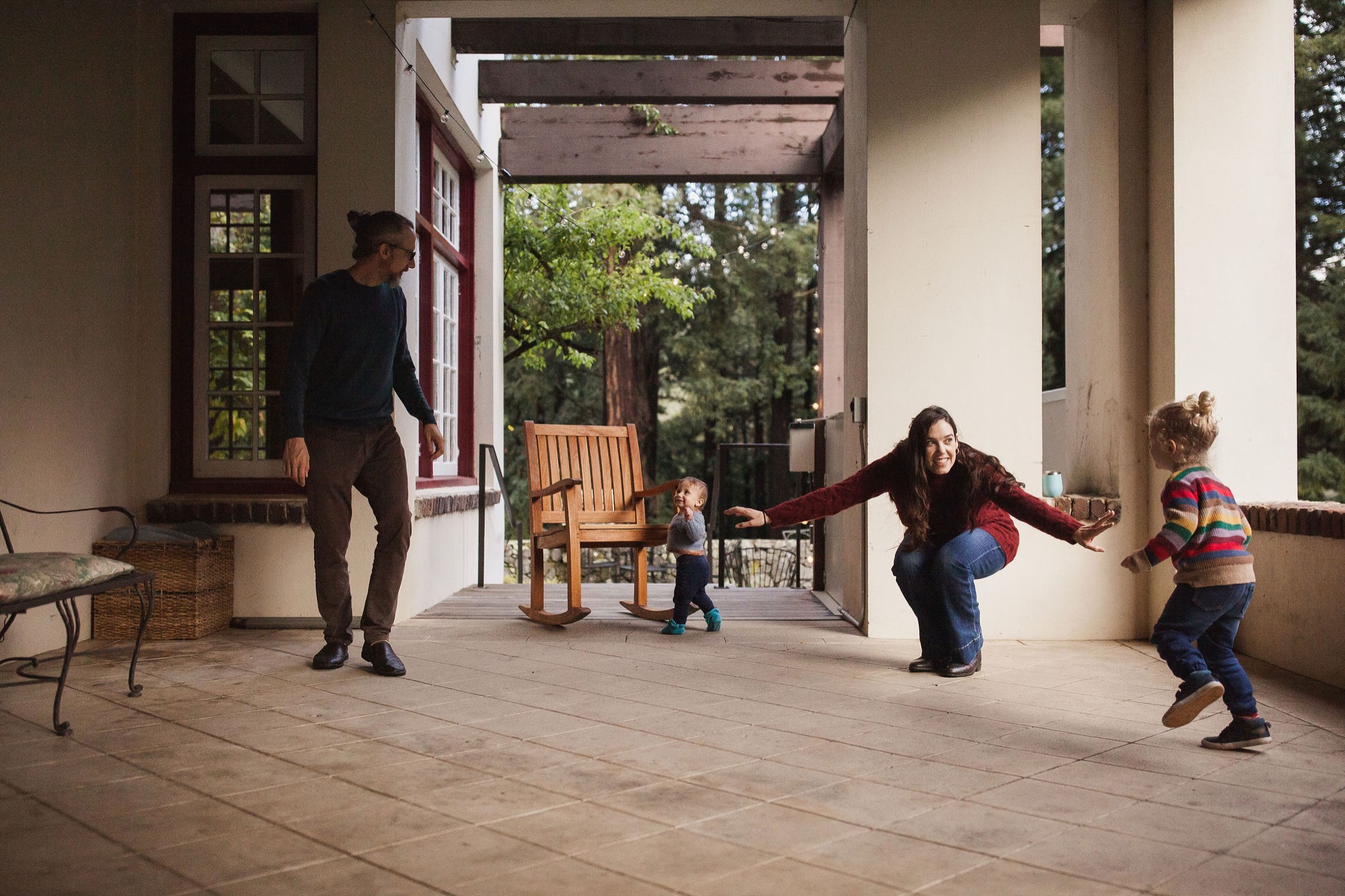 A woman crouches with arms spread wide as two young children run toward her on a sun-dappled covered porch, while a man watches from the left