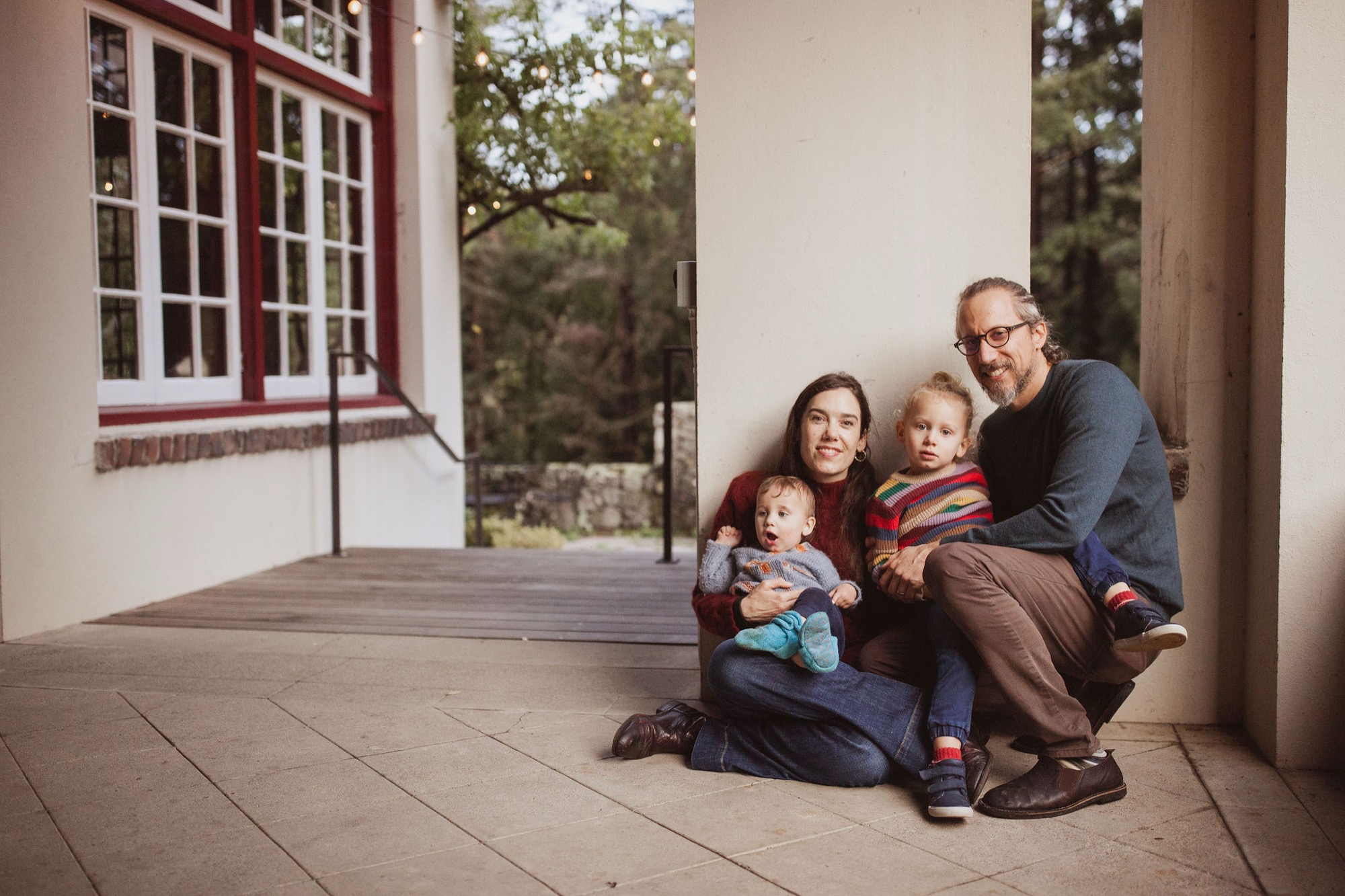 A family of four crouches together on a tiled porch, the children in colorful striped sweaters while soft natural light frames their genuine smiles against white walls