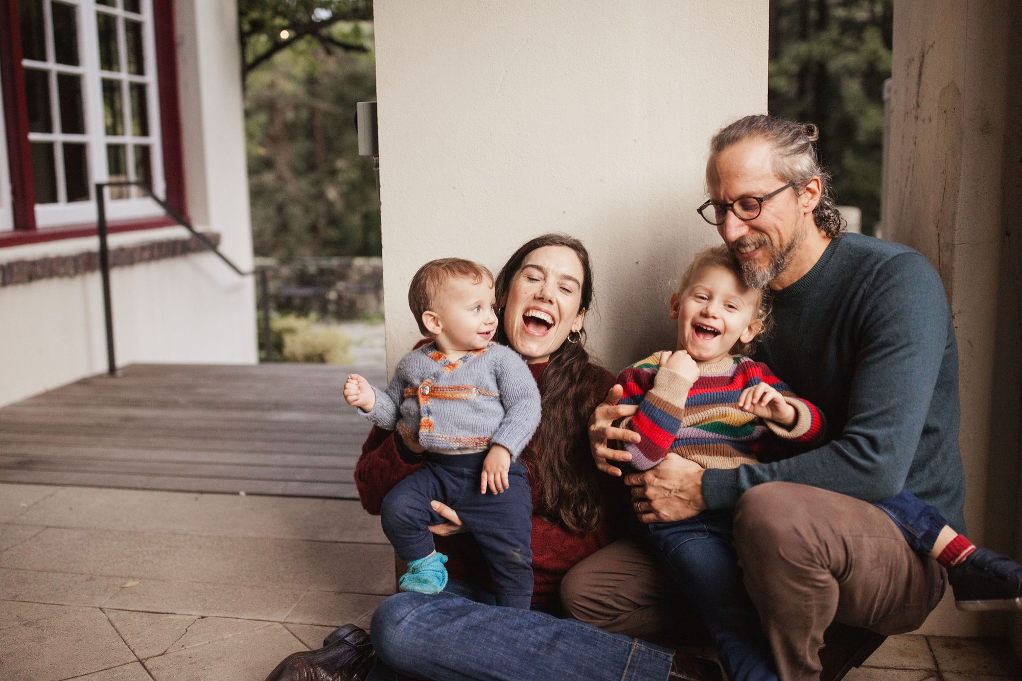 A family of four crouches together on a sunny patio, their genuine laughter and close physical contact creating an intimate moment against a white wall