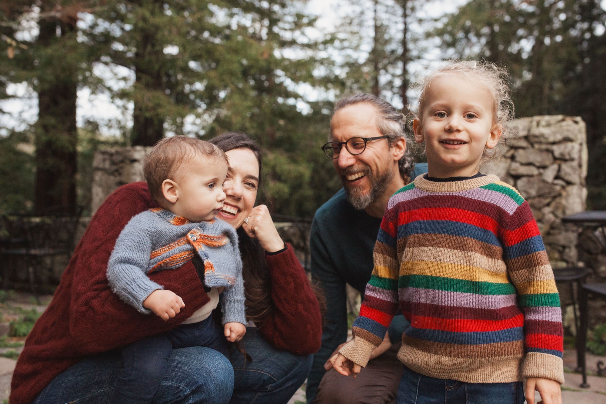 A family of four laughs together beside weathered stone walls, with the father's hand on his daughter's shoulder while autumnal trees frame the background