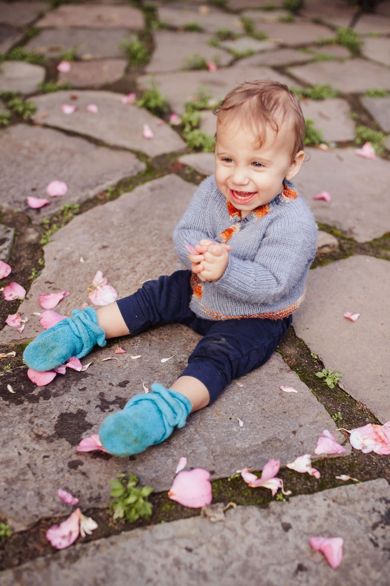 Toddler sits laughing on moss-covered pavement scattered with pink flower petals, wearing blue mittens and gray cardigan