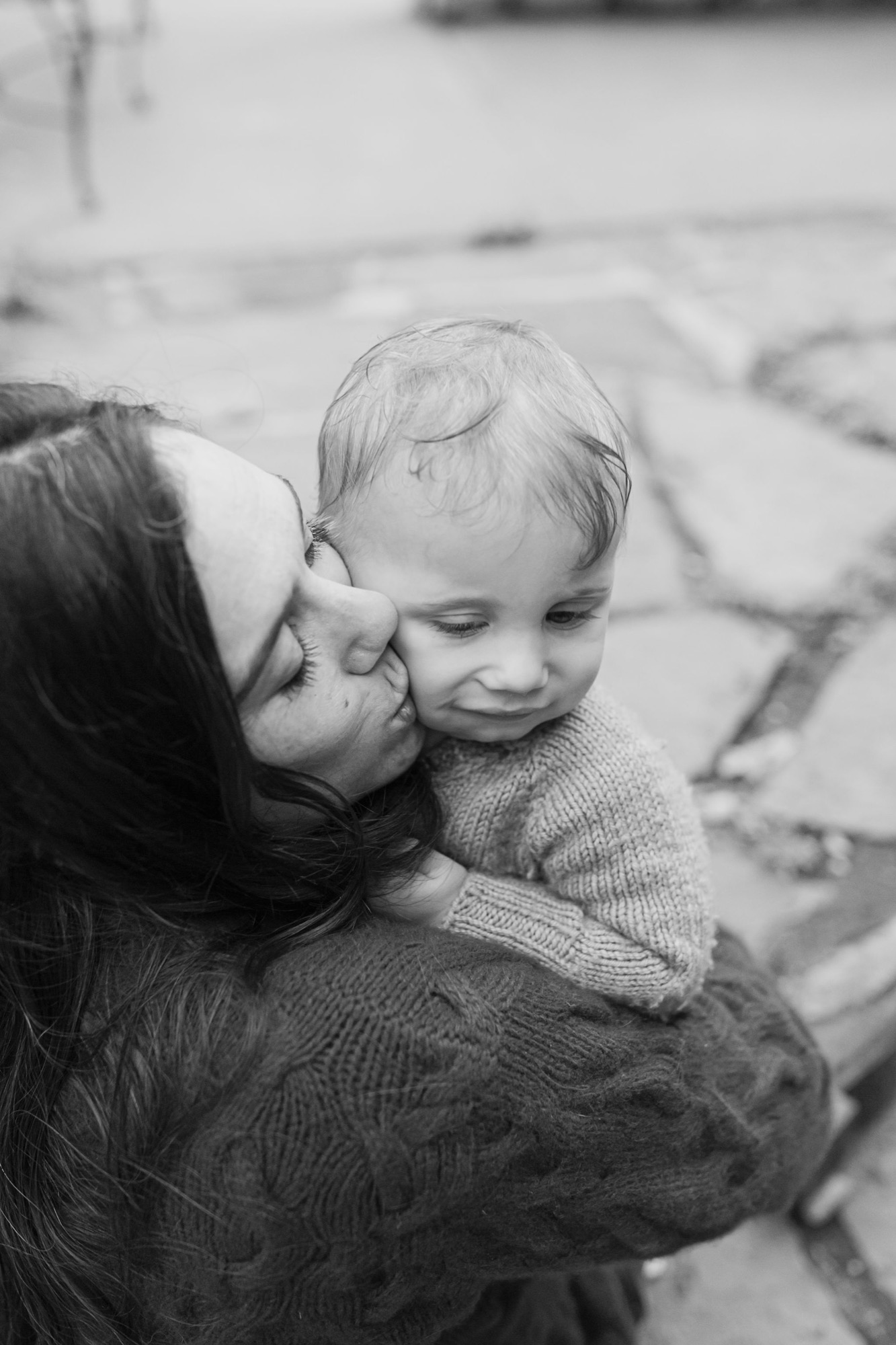 A mother kisses her toddler's cheek while holding him, both wearing knit sweaters in soft natural light
