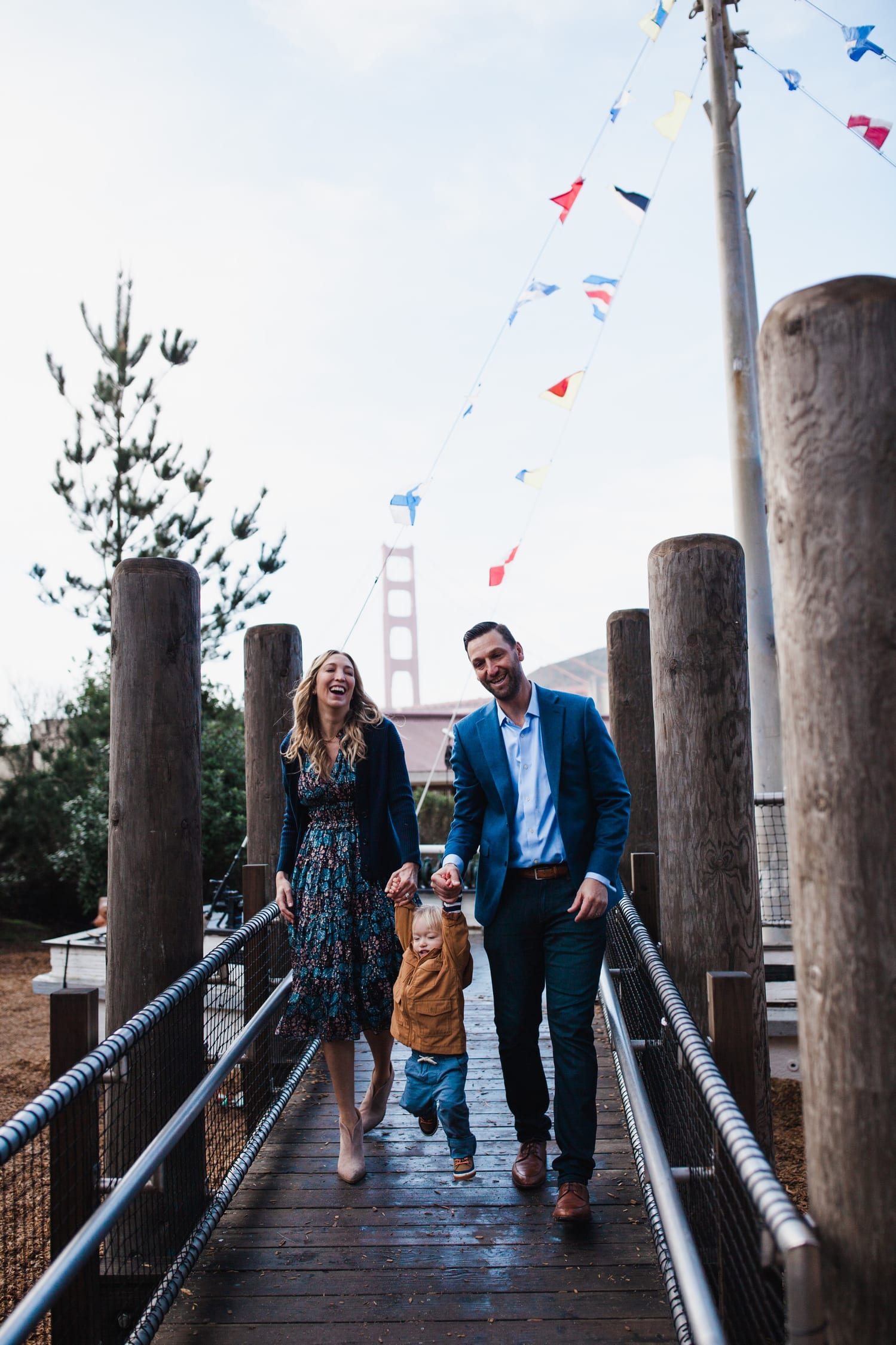 Parents swing toddler by the hands on a boardwalk, Golden Gate Bridge behind