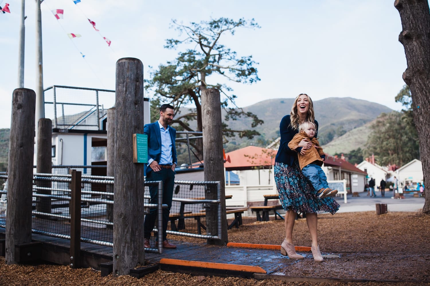 Mom laughs and swings her toddler at a playground while dad watches