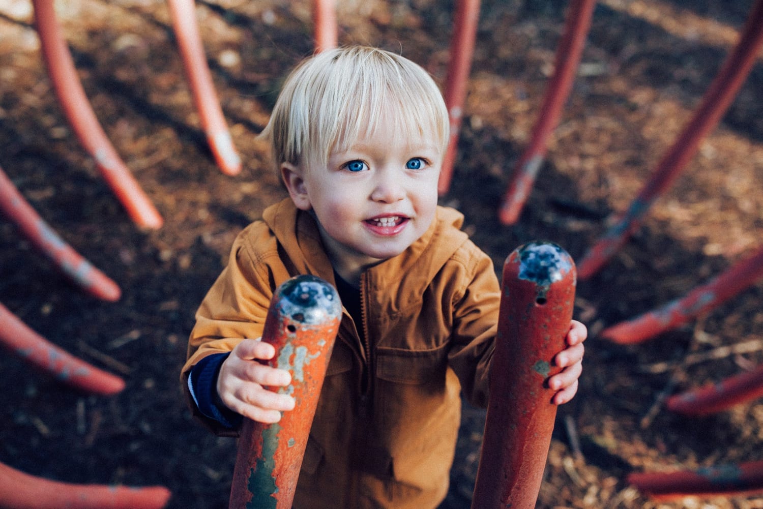 Toddler grips red playground climbing bars and smiles up at the camera