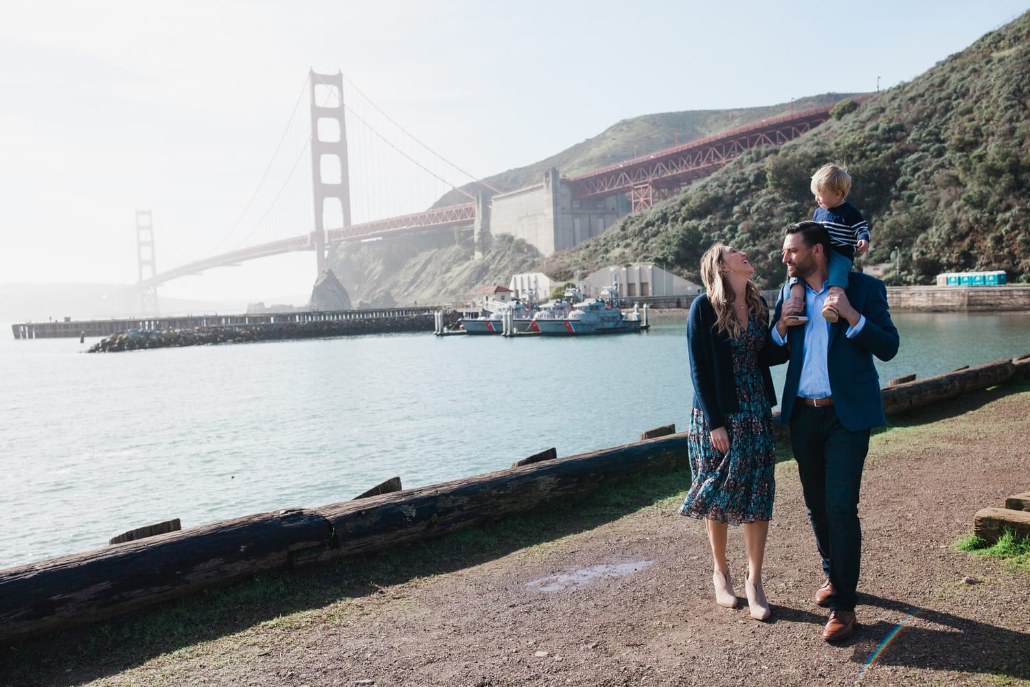 Mom looks up at toddler on dad's shoulders, Golden Gate Bridge in the distance
