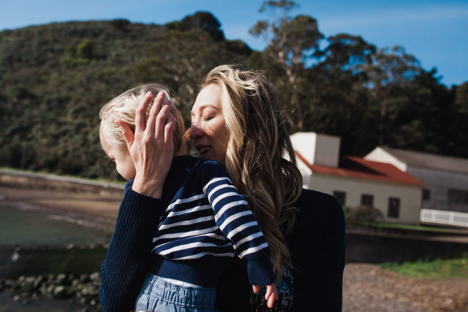 Mom hugs her toddler close, cradling his head, hillside and building behind