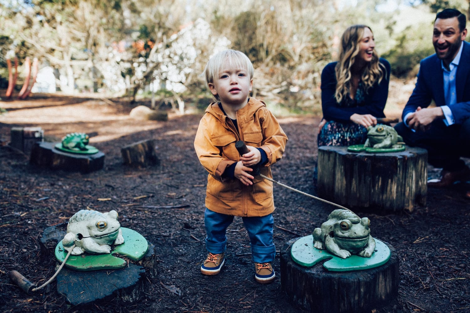 Toddler plays with wooden frog sculptures while his parents laugh behind him
