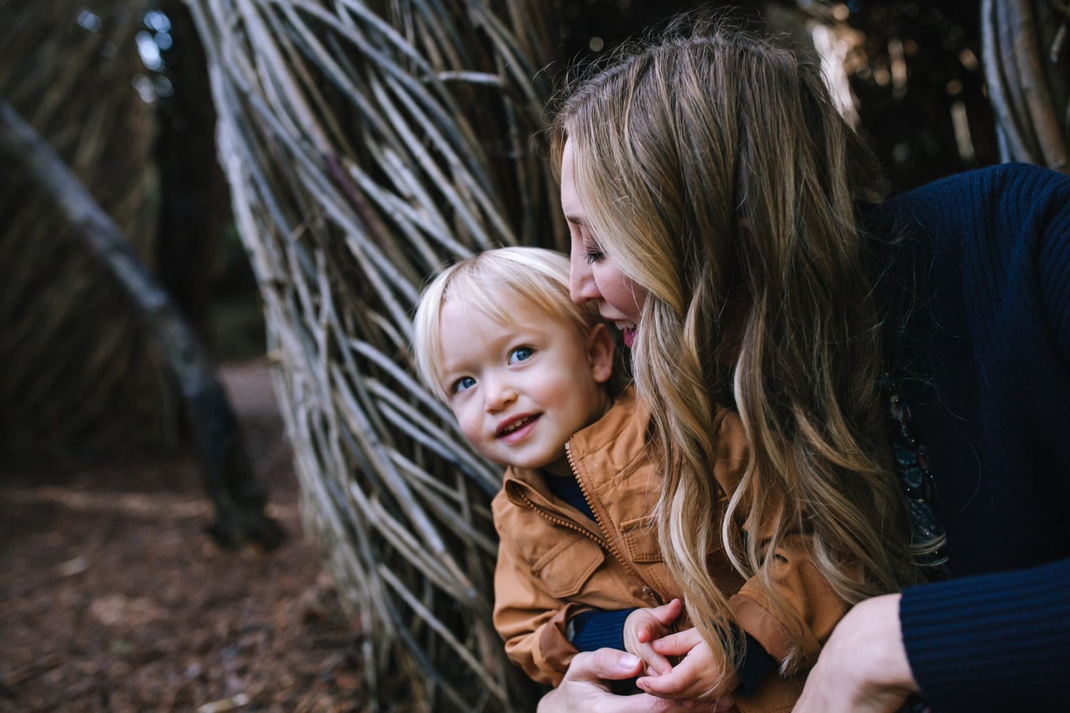 Mom cuddles her smiling toddler beside a woven branch structure