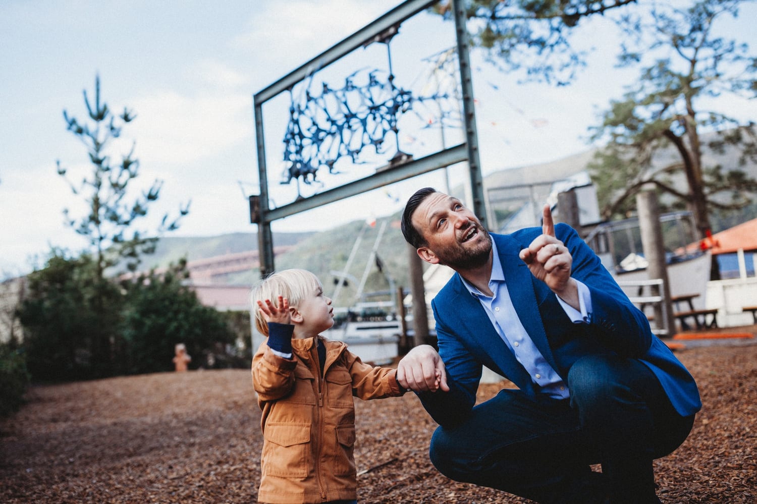 Dad crouches beside his toddler and points up at a playground sculpture