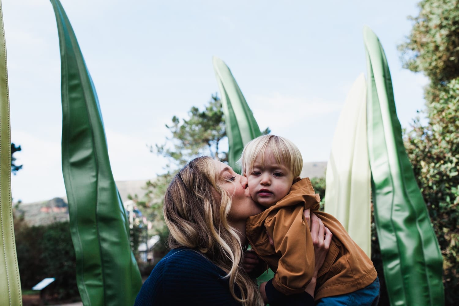 Mom kisses her toddler's cheek among giant green leaf sculptures