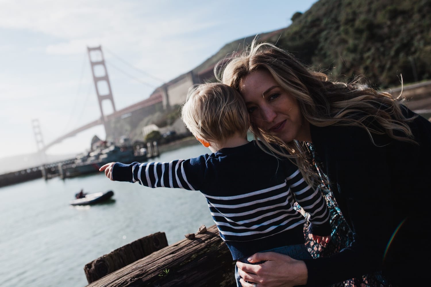 Mom holds her toddler as he points across the water, Golden Gate Bridge behind