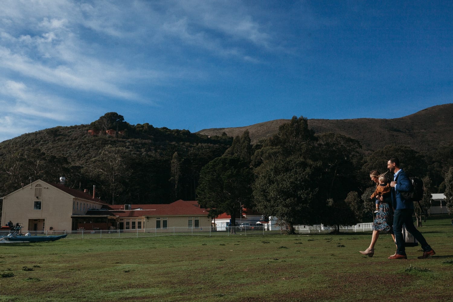Family walks across a grassy field past historic buildings below wooded hills