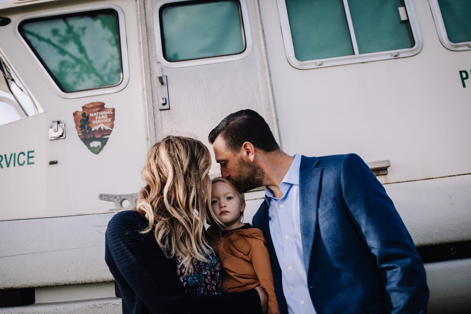 Dad kisses toddler's head while mom holds him beside a National Park Service boat