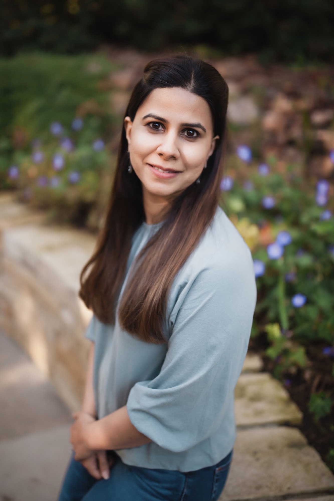 Woman in light blue top smiles warmly toward camera, positioned beside blooming blue flowers in soft natural garden light